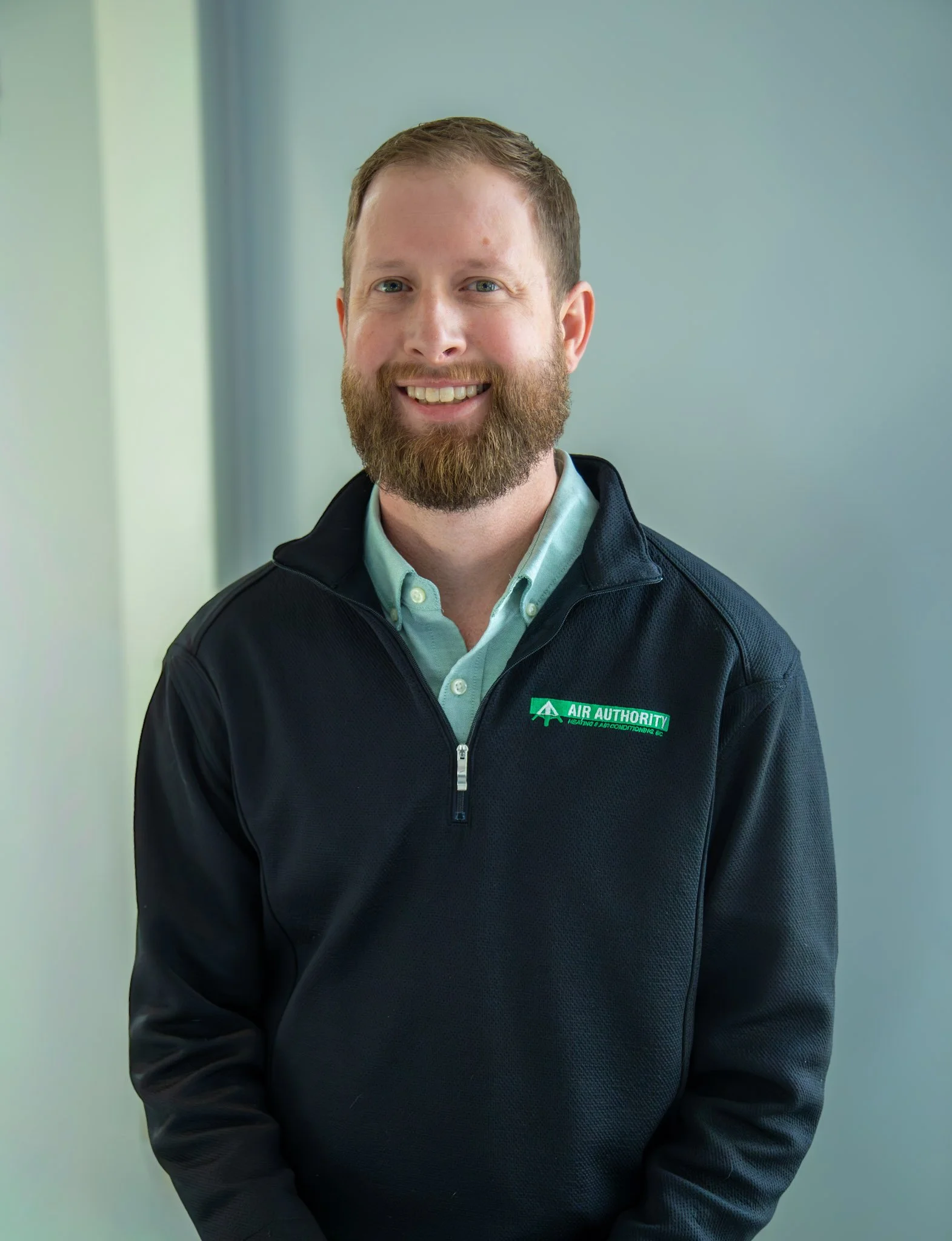 A professional man with a beard and short hair, wearing a black jacket with a logo that reads 'Air Authority Heating & Air Conditioning.' He is smiling and standing in an indoor setting with a plain wall background.
