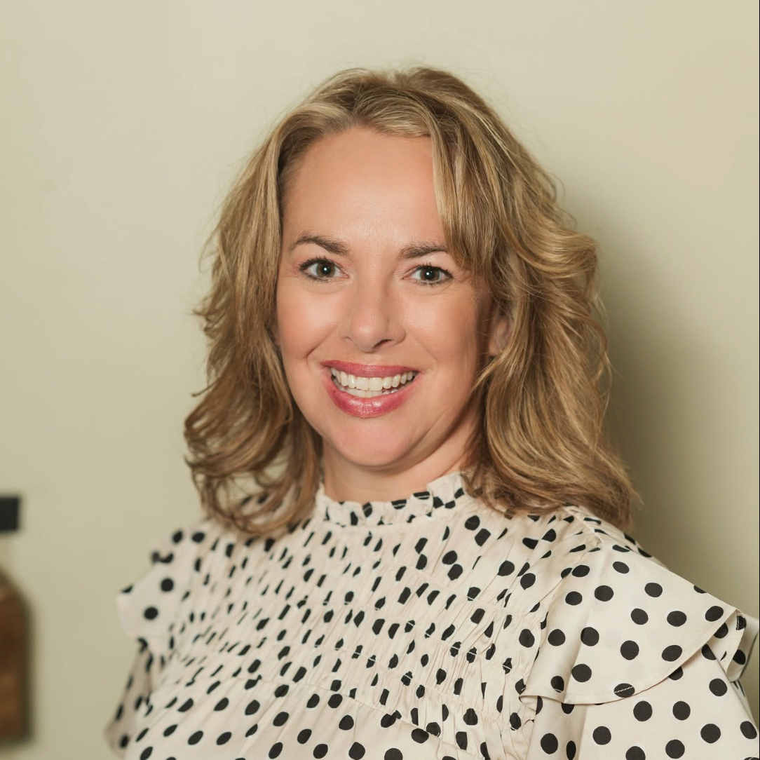 A woman with blonde hair smiling at the camera, wearing a white blouse with black polka dots, standing against a light-colored wall.