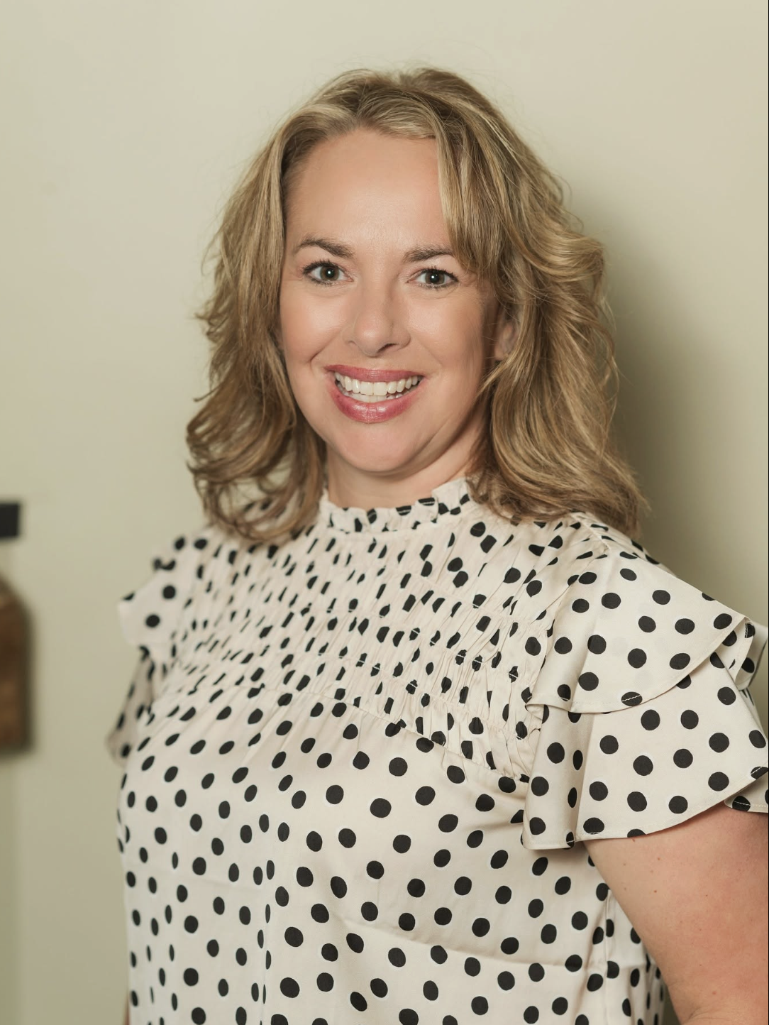 A woman with shoulder-length blonde hair smiling, wearing a white blouse with black polka dots, in front of a plain, light-colored wall.
