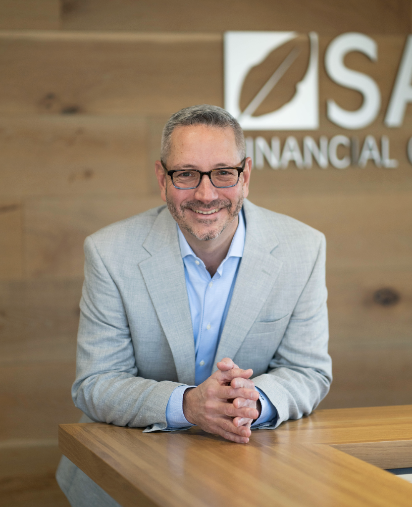 A man with glasses, wearing a light gray suit and blue shirt, sitting at a wooden desk in an office with a wooden wall and logo that reads 'SAGE Financial' behind him.
