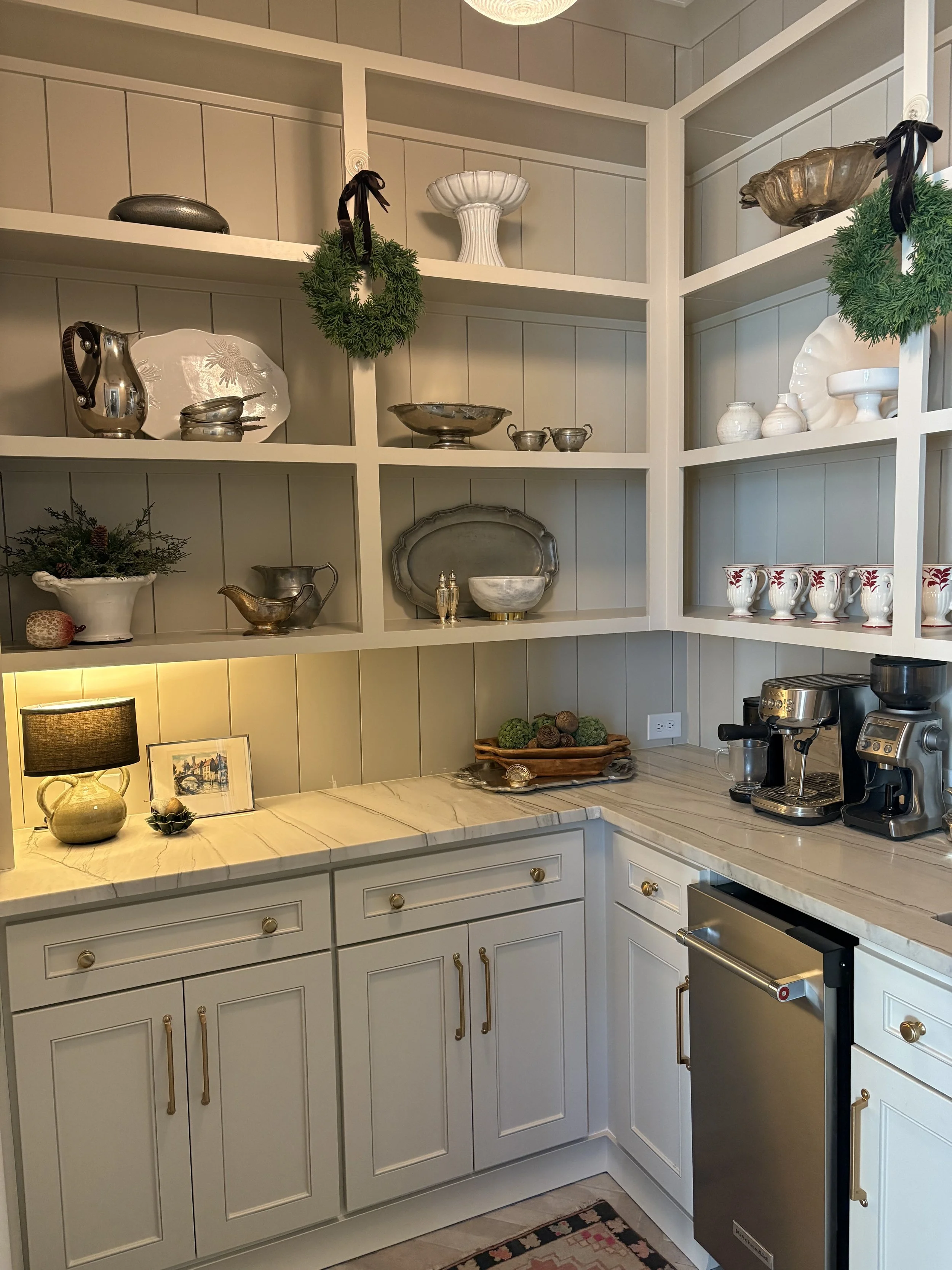A kitchen with white cabinets and marble countertops. Open shelving displays silver and white decorative items, including vases, bowls, and dishes. A coffee machine and other appliances are on the counter. Green holiday wreaths hang on the shelves.