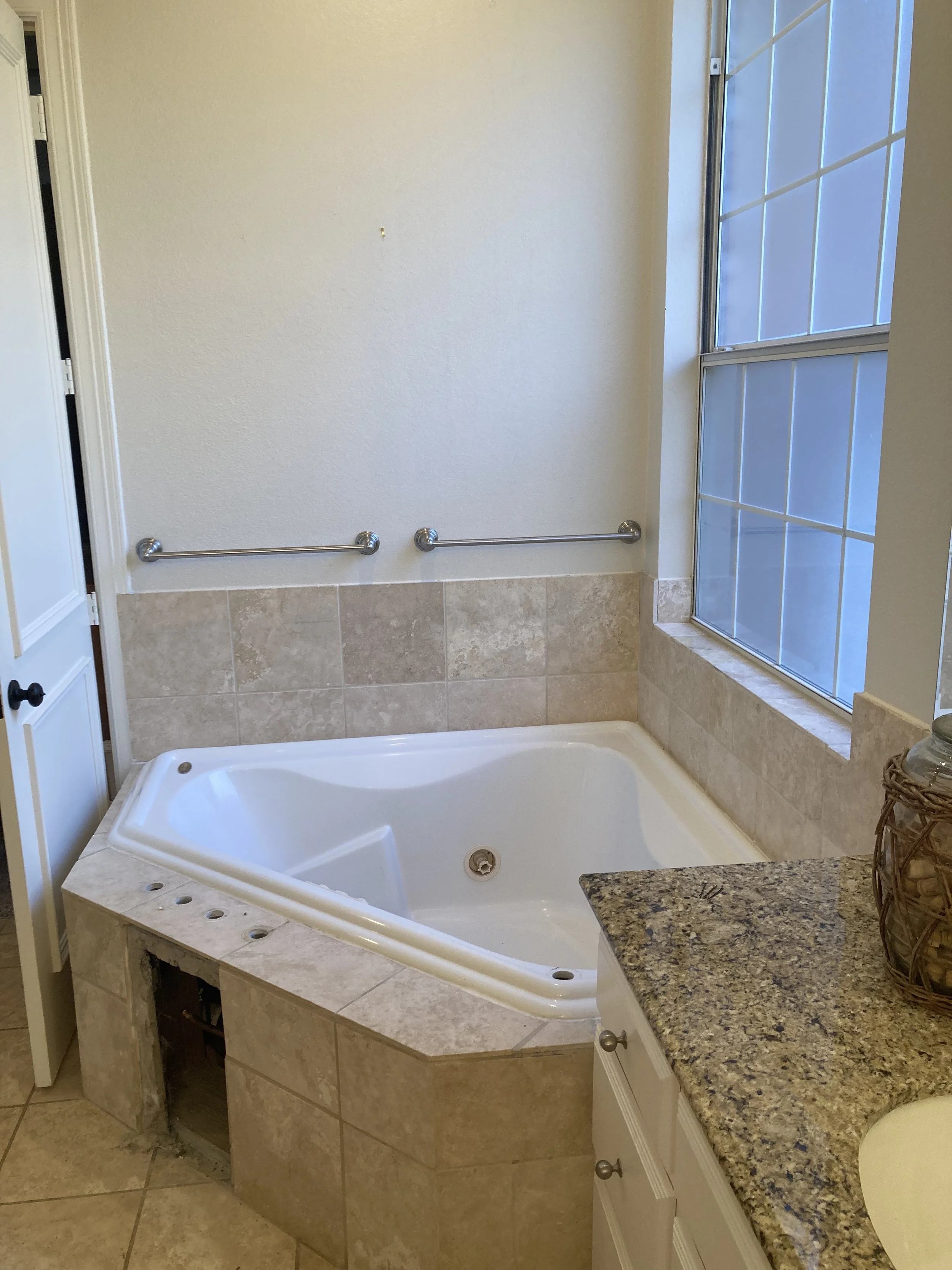 Bathroom with a large whirlpool bathtub, beige tile surround, two metal grab bars on the wall, and a granite countertop with a basket of decorative items, next to a large window with grid pattern glass.