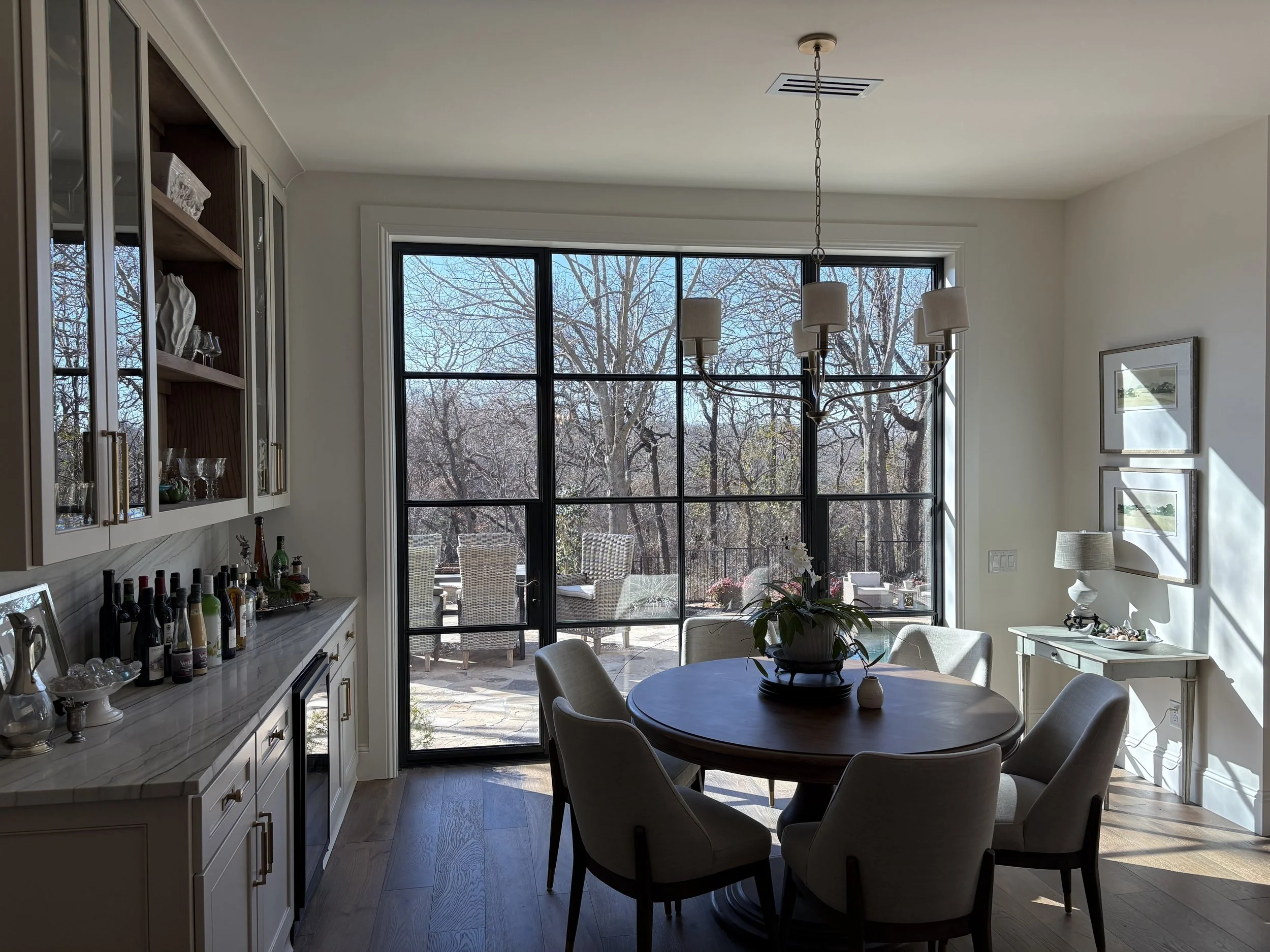 A dining area with a round table and six chairs, located near large windows showing a view of trees and outdoor furniture. A chandelier hangs above the table, and a side table with framed pictures and a lamp is visible on the right. The room has a light color palette with white walls and cabinetry.