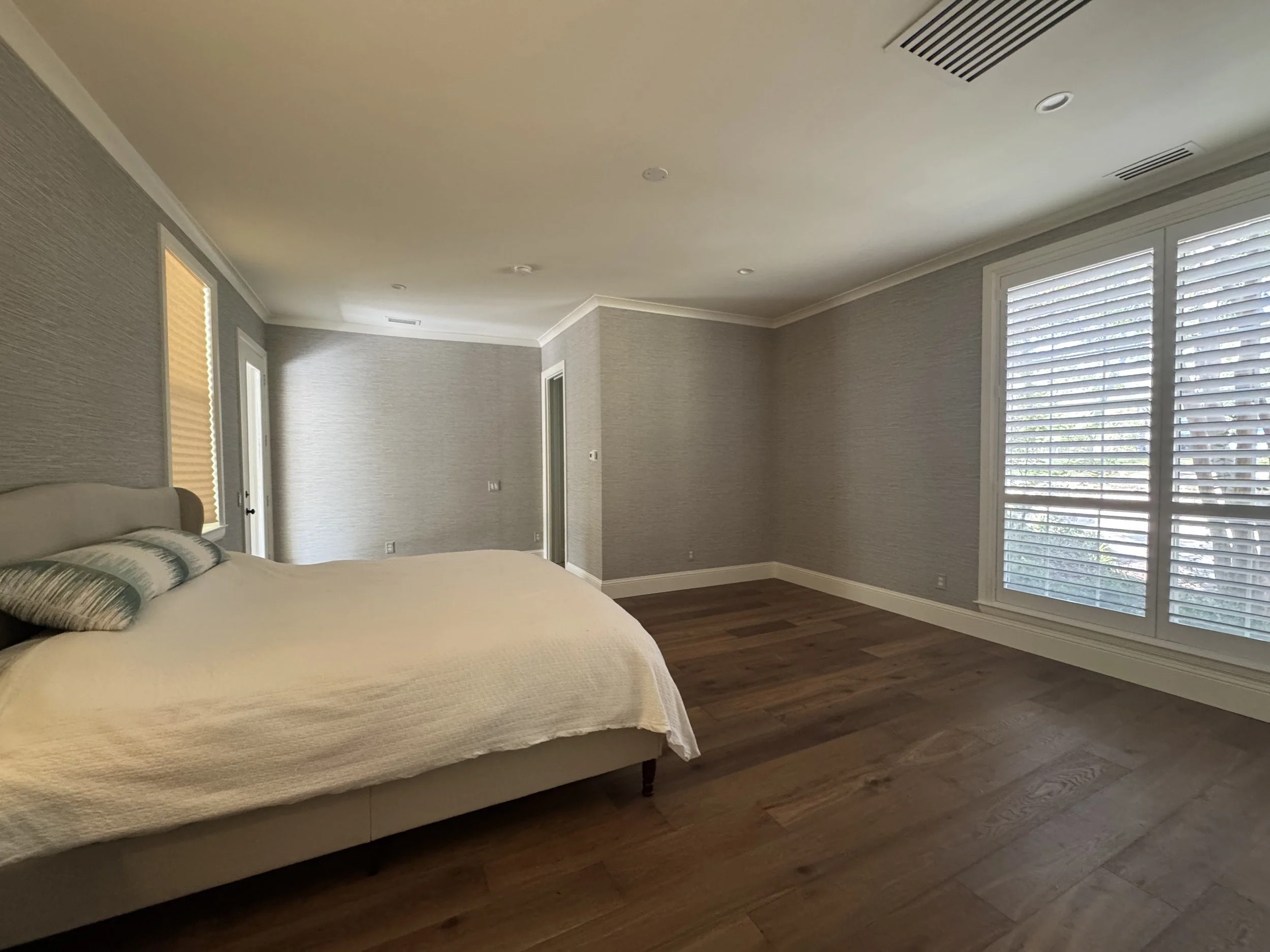 A bedroom with light-colored walls, a bed with a white quilt and pillows, windows with white blinds, wood flooring, and some recessed lighting in the ceiling.