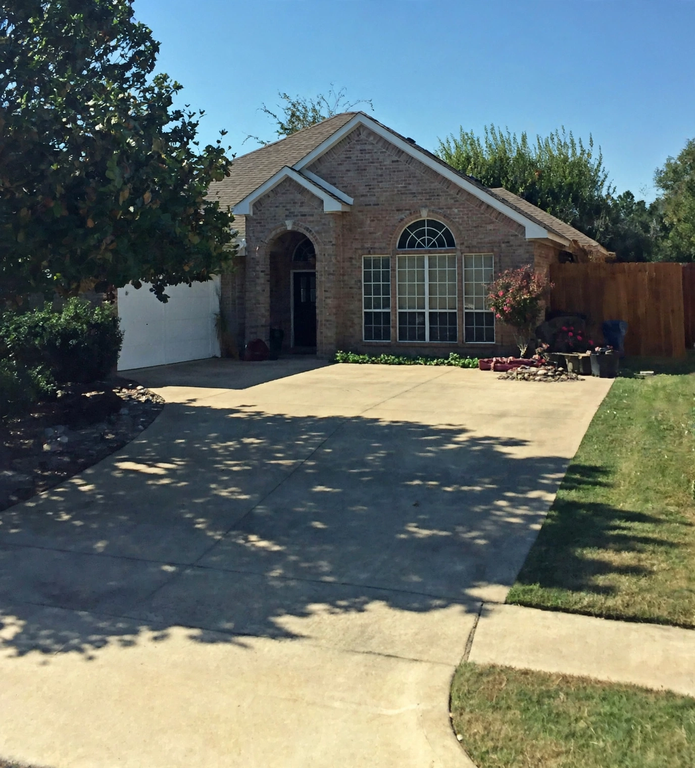 A brick house with large front windows, a garage on the left, and a driveway in front. There are trees and plants around the house, a wooden fence on the right, and a clear blue sky above.