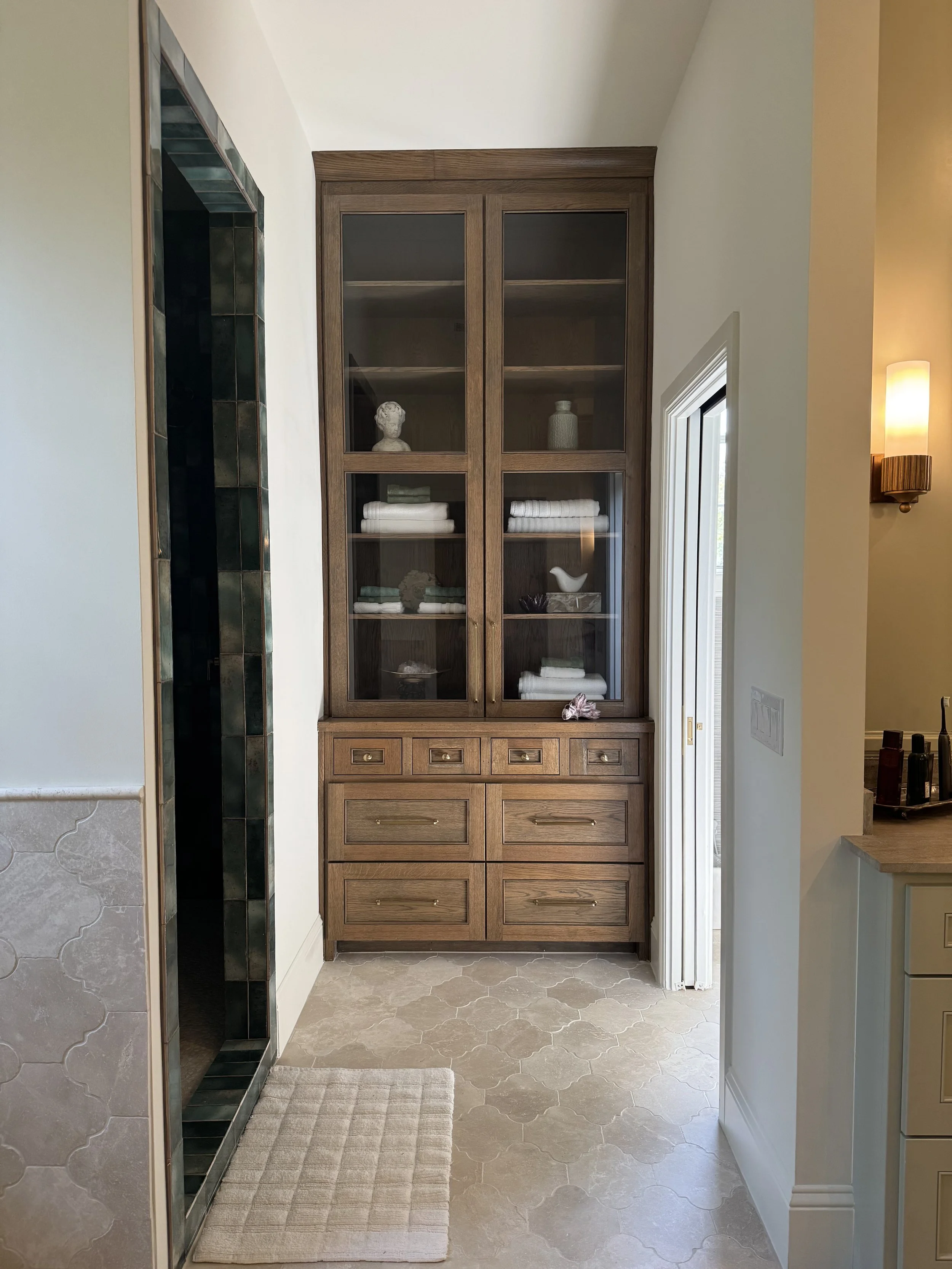 A hallway with a built-in wooden cabinet with glass doors, containing neatly folded towels and decorative items, next to a wall with a light on.