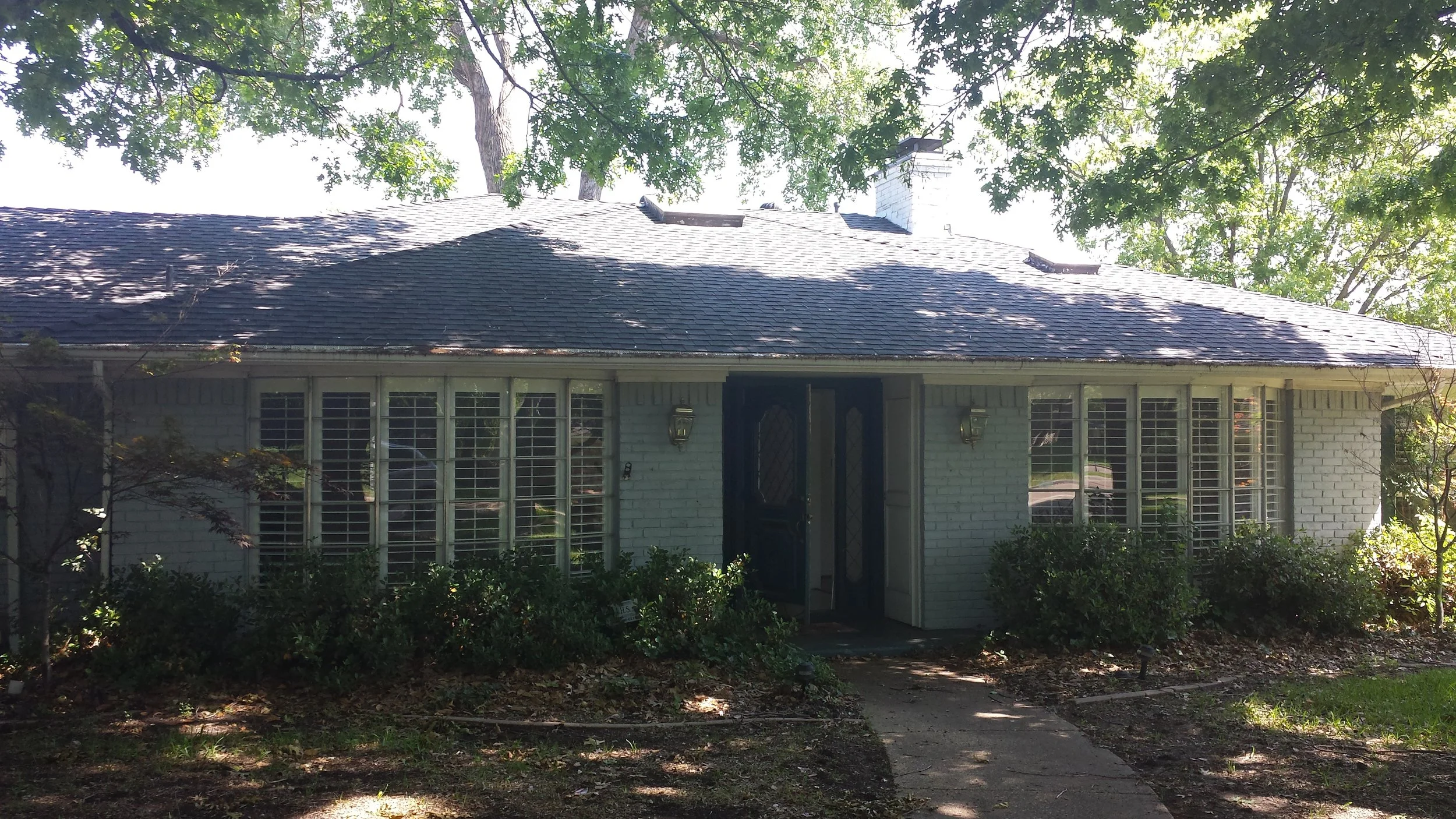 A house with a brick exterior and large window panes, a black front door, and a shingled roof with a chimney, surrounded by trees and greenery.