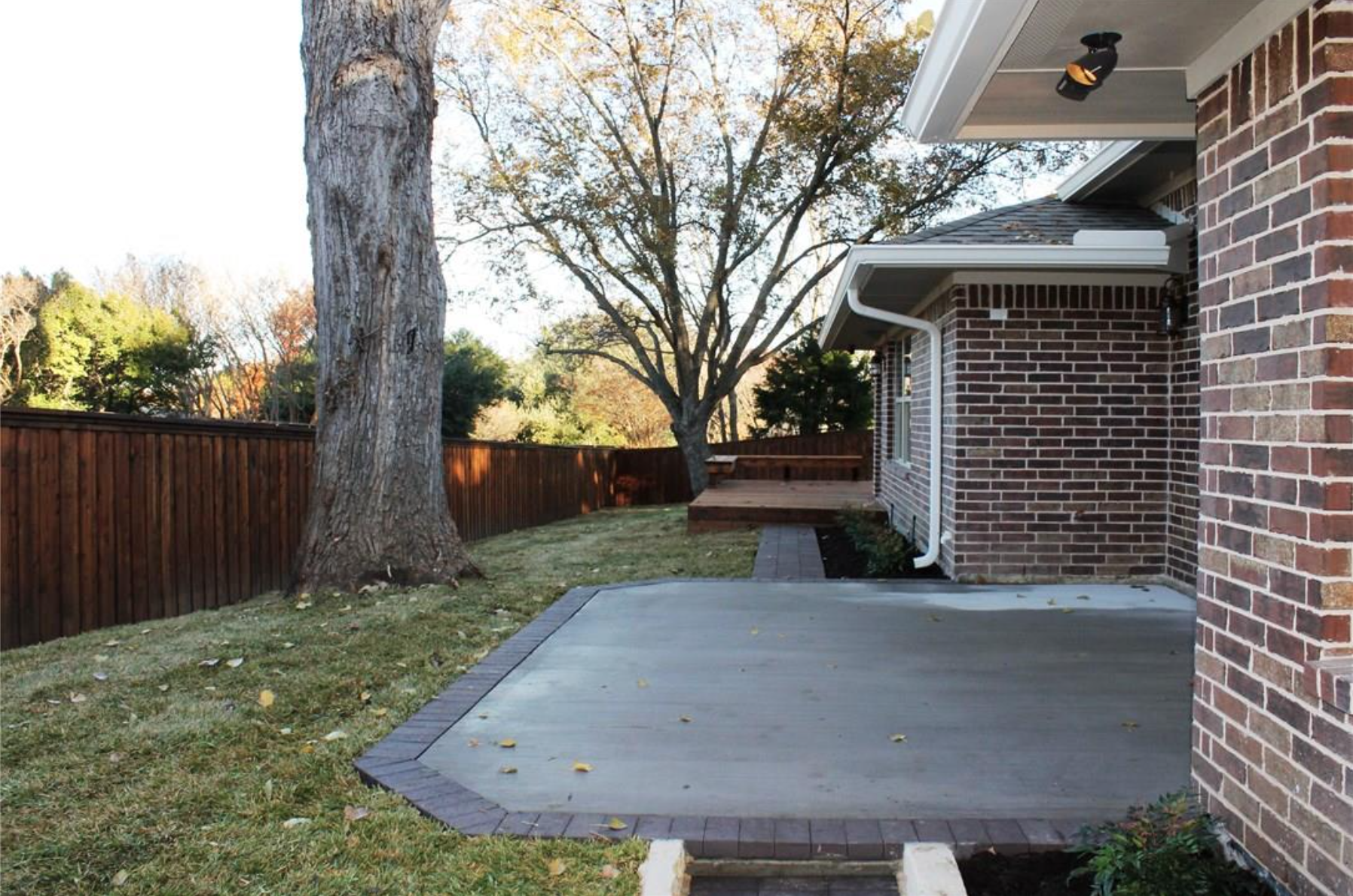 A backyard with a concrete patio edged with bricks, a grassy area with fallen leaves, a tall tree with no leaves, and a wooden fence. Part of a brick house with a roof fixture is visible on the right.