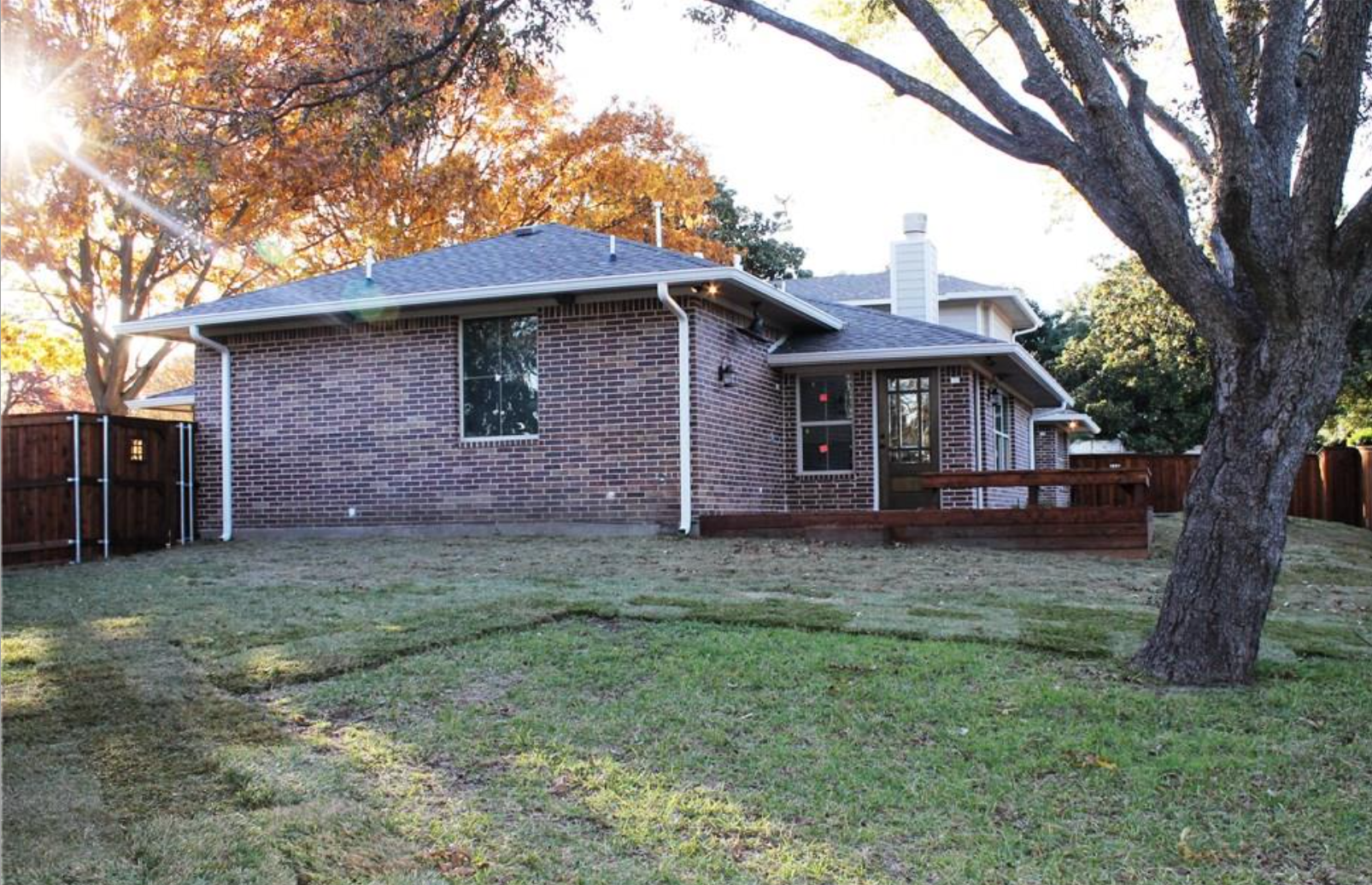 Backyard of a brick house with a wooden deck, surrounded by a wooden fence, large trees, and a grassy lawn.