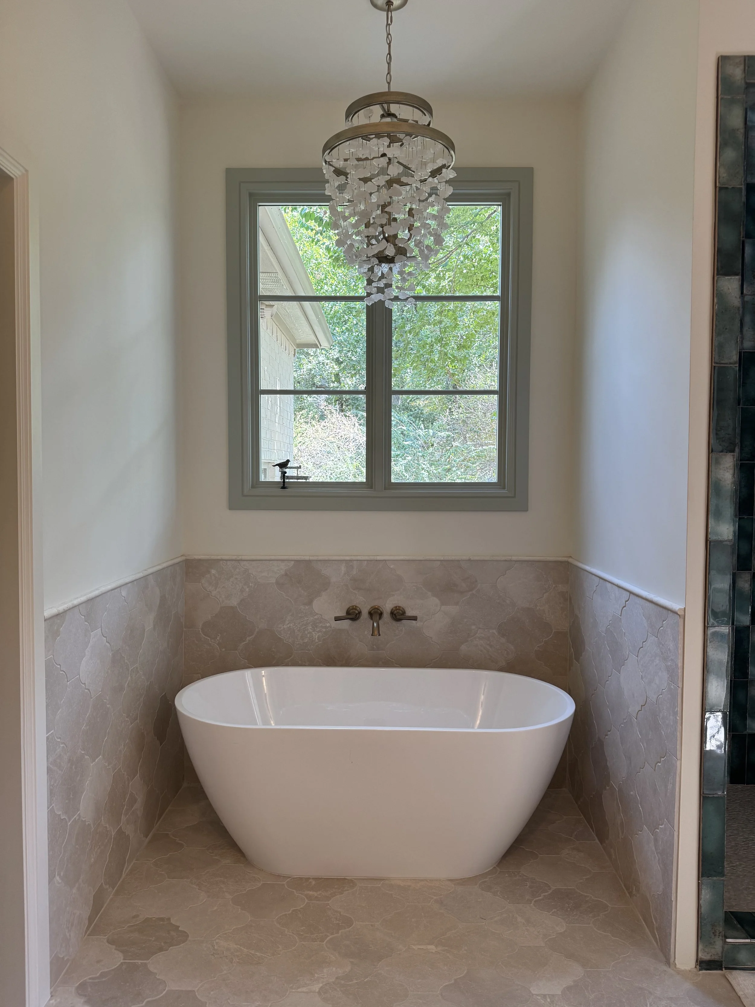 A bathroom with a freestanding white bathtub below a window, with a chandelier hanging above and a window view of green trees.