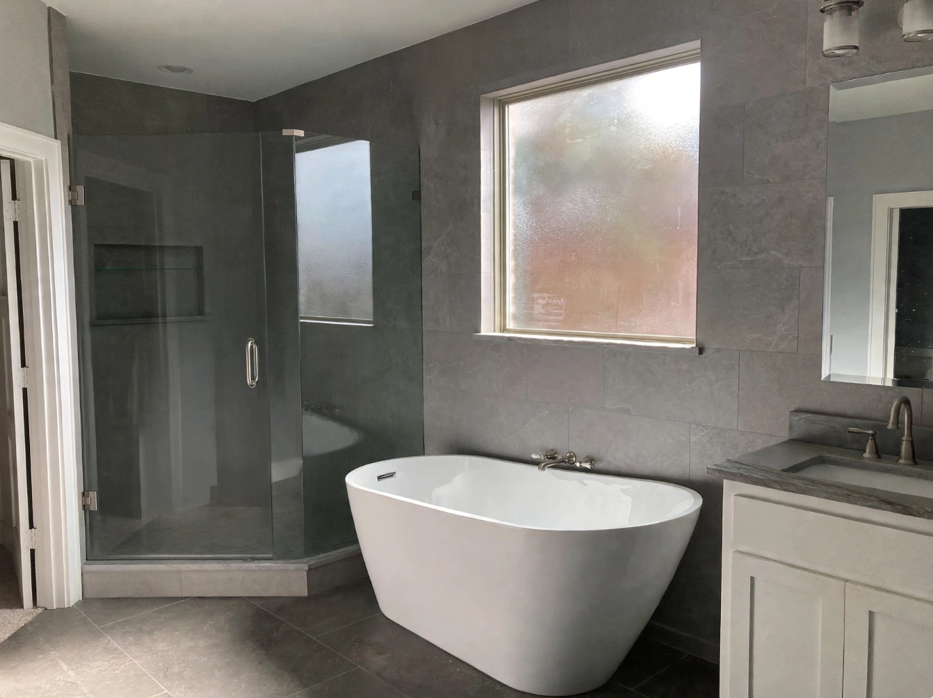Modern bathroom with a freestanding bathtub, glass-enclosed shower, gray tiled walls and floors, and a small vanity with a gray countertop and mirror.