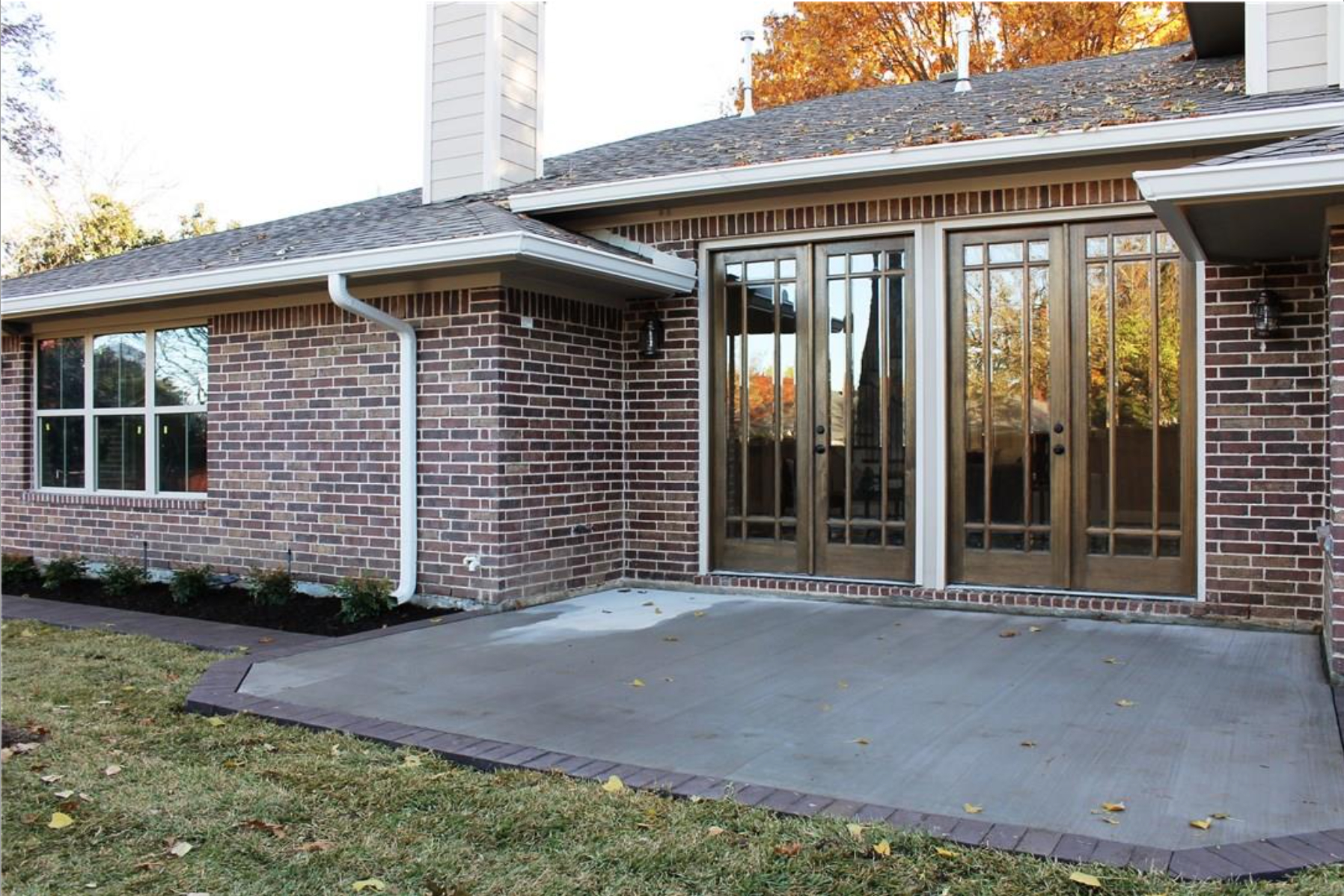Backyard patio with concrete flooring, bordered by brick and a black strip, adjacent to a house with brick exterior and large wooden glass doors, surrounded by grass, small plants, and trees with fall leaves.