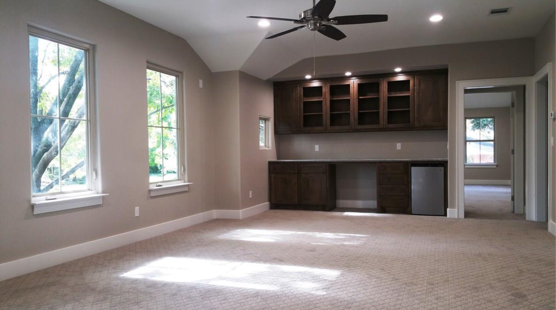 An empty room with beige walls, three large windows on the left, a ceiling fan, built-in wooden cabinets with glass doors, and a small refrigerator under the cabinets. There's a doorway to another room on the right.