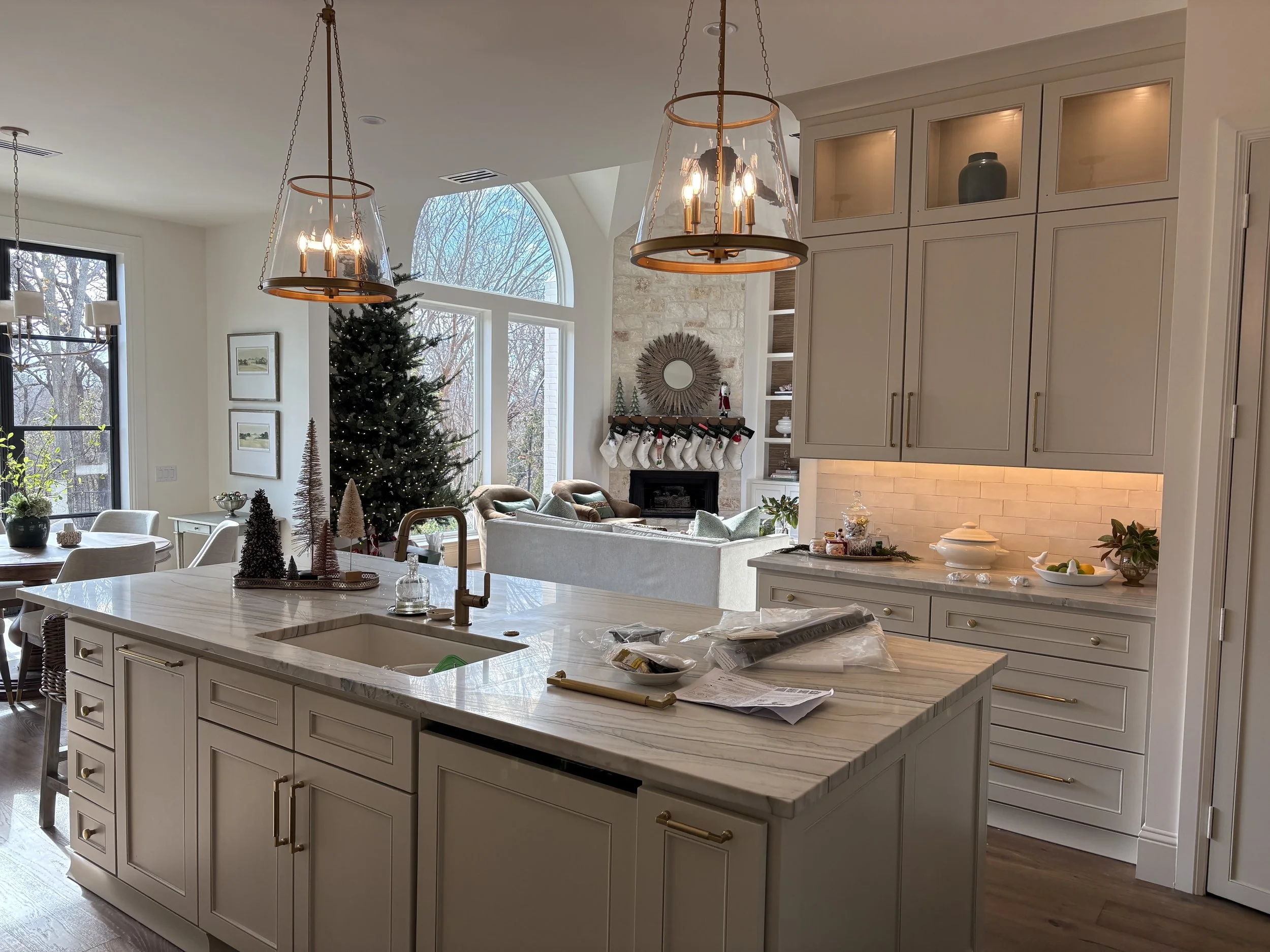 A spacious kitchen with a large white marble island, modern pendant lighting, and cabinetry with gold hardware. In the background, there's a cozy living area with a decorated Christmas tree and stockings hanging from a fireplace.