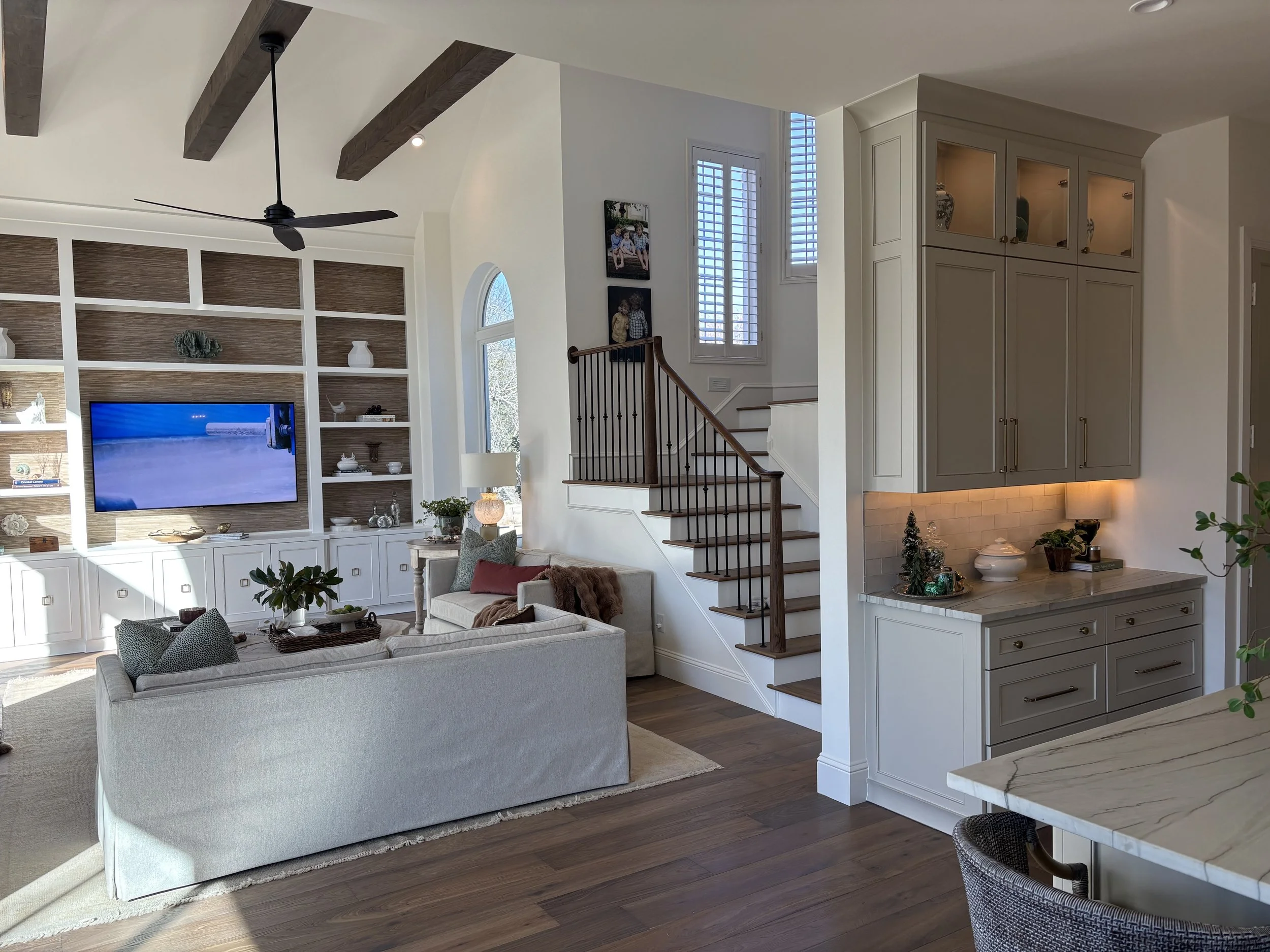Modern living room with white walls, a built-in entertainment center with a large flat-screen TV, and a staircase with a dark wood handrail. There is a white sofa with decorative pillows, a side table with a lamp, and large windows with plantation shutters allowing natural light. The ceiling has wooden beams and a black ceiling fan.