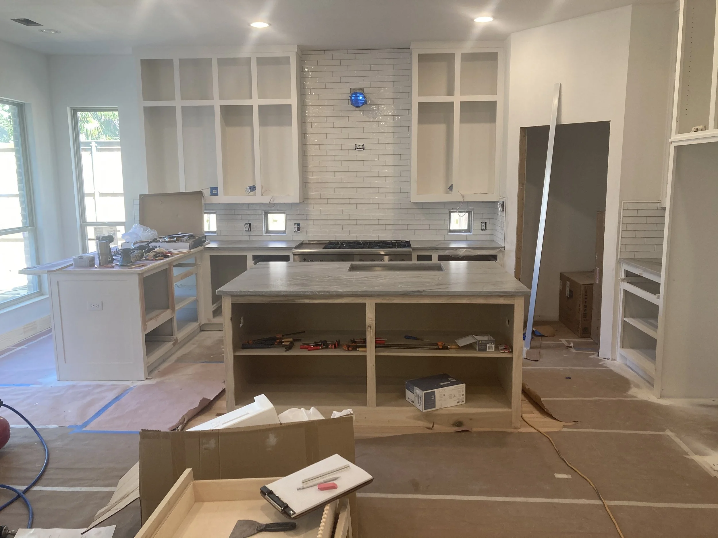 Interior of a kitchen under renovation with white cabinets, a brick backsplash, and construction tools and materials on the counters and floor.