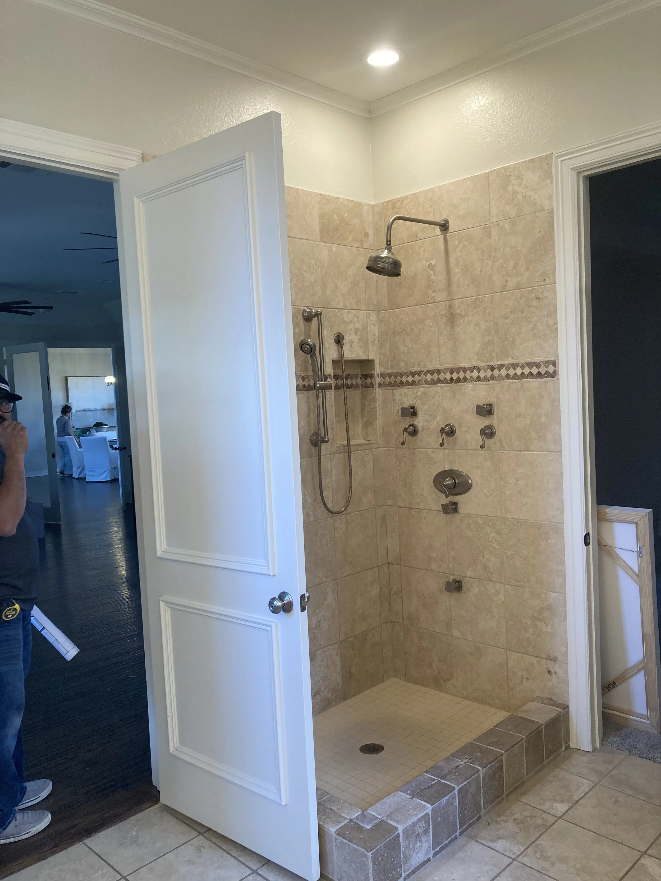 Bathroom shower area with beige tiles, a built-in shower niche, a rainfall showerhead, multiple water control handles, and a step with a brick border. The shower is open with a partially open white door.