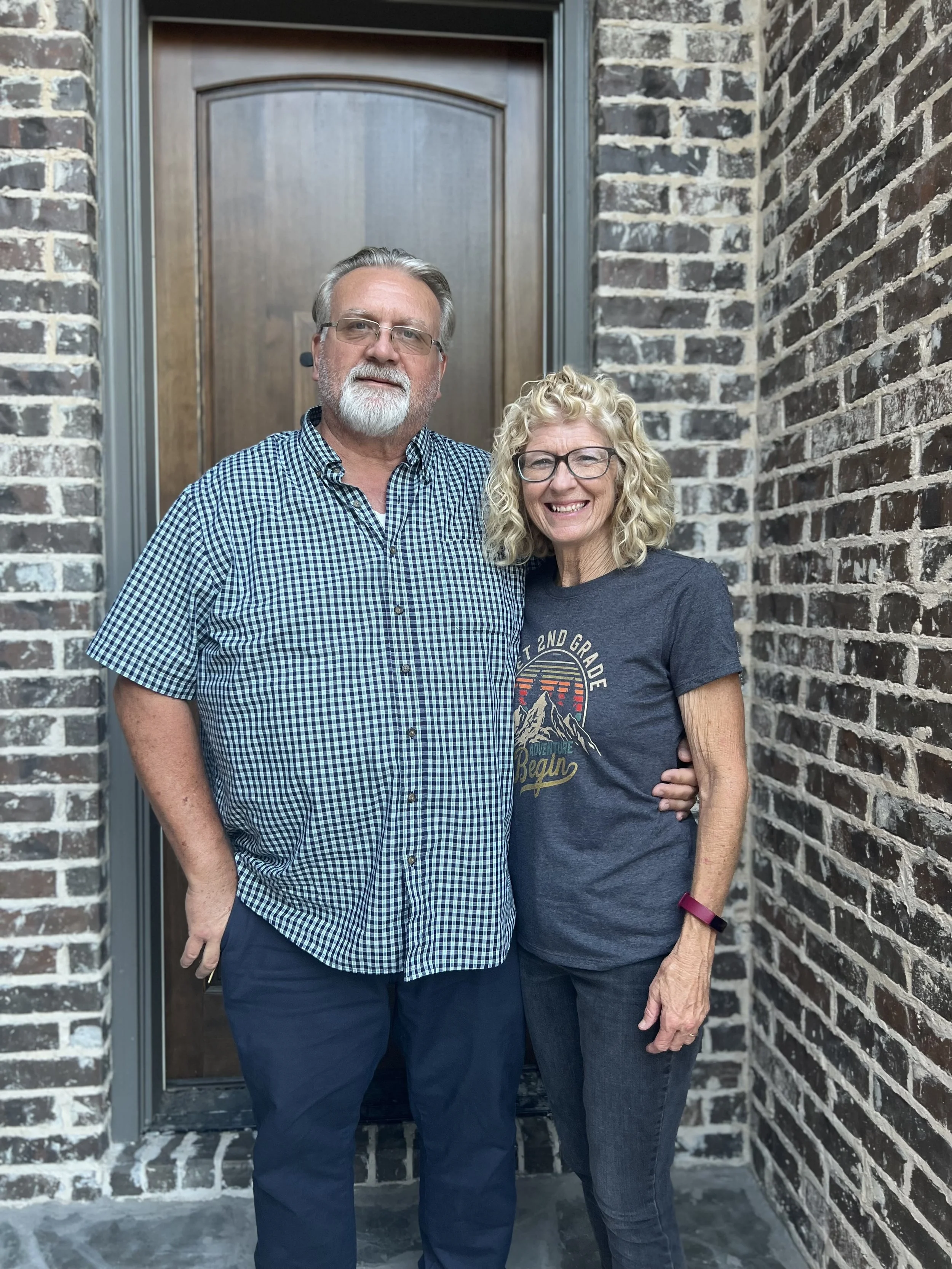 A man and woman standing close together in front of a brick wall and a wooden door, smiling for the photo. The man is wearing glasses and a checkered shirt, and the woman is wearing glasses and a dark T-shirt with a graphic print.