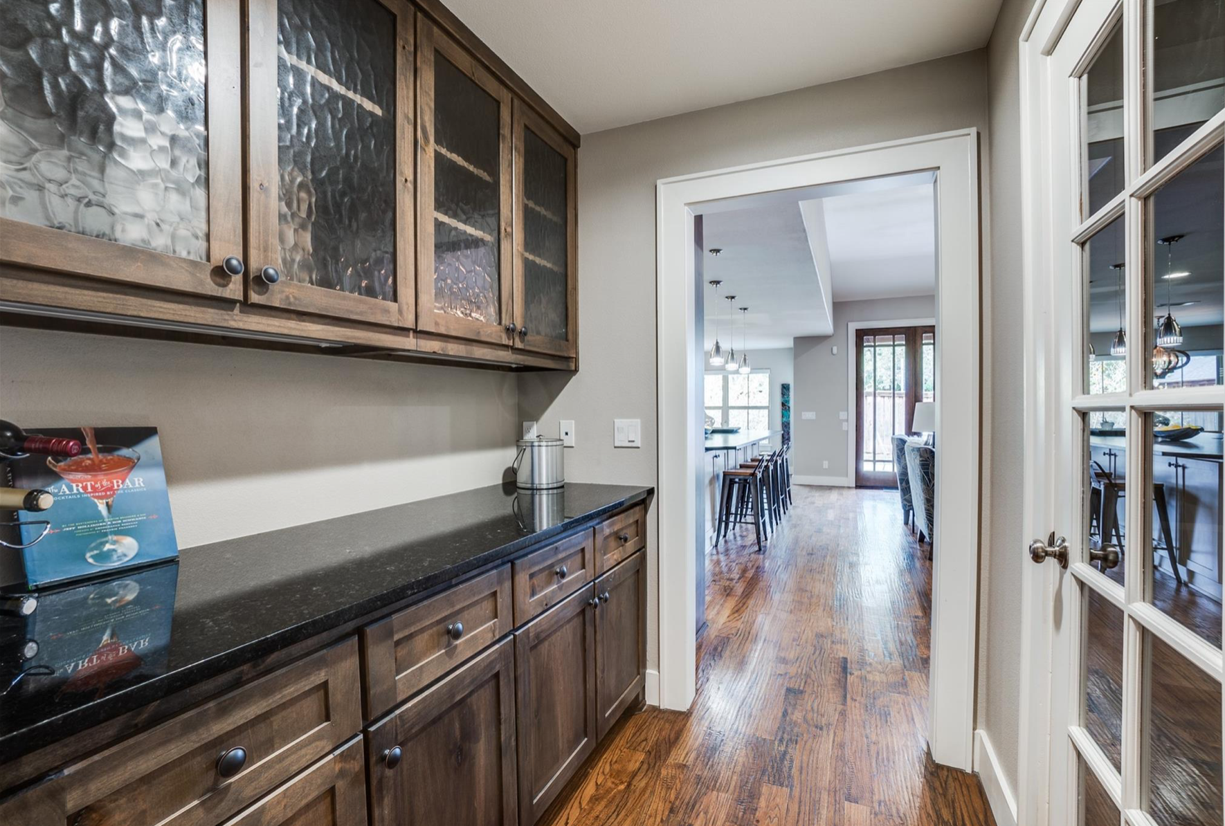 View from a kitchen showing dark wooden cabinets with textured glass doors, black countertops, and a wooden floor. A doorway leads to a bright dining area with a breakfast bar, bar stools, and a living room with large windows and a door to the outside.