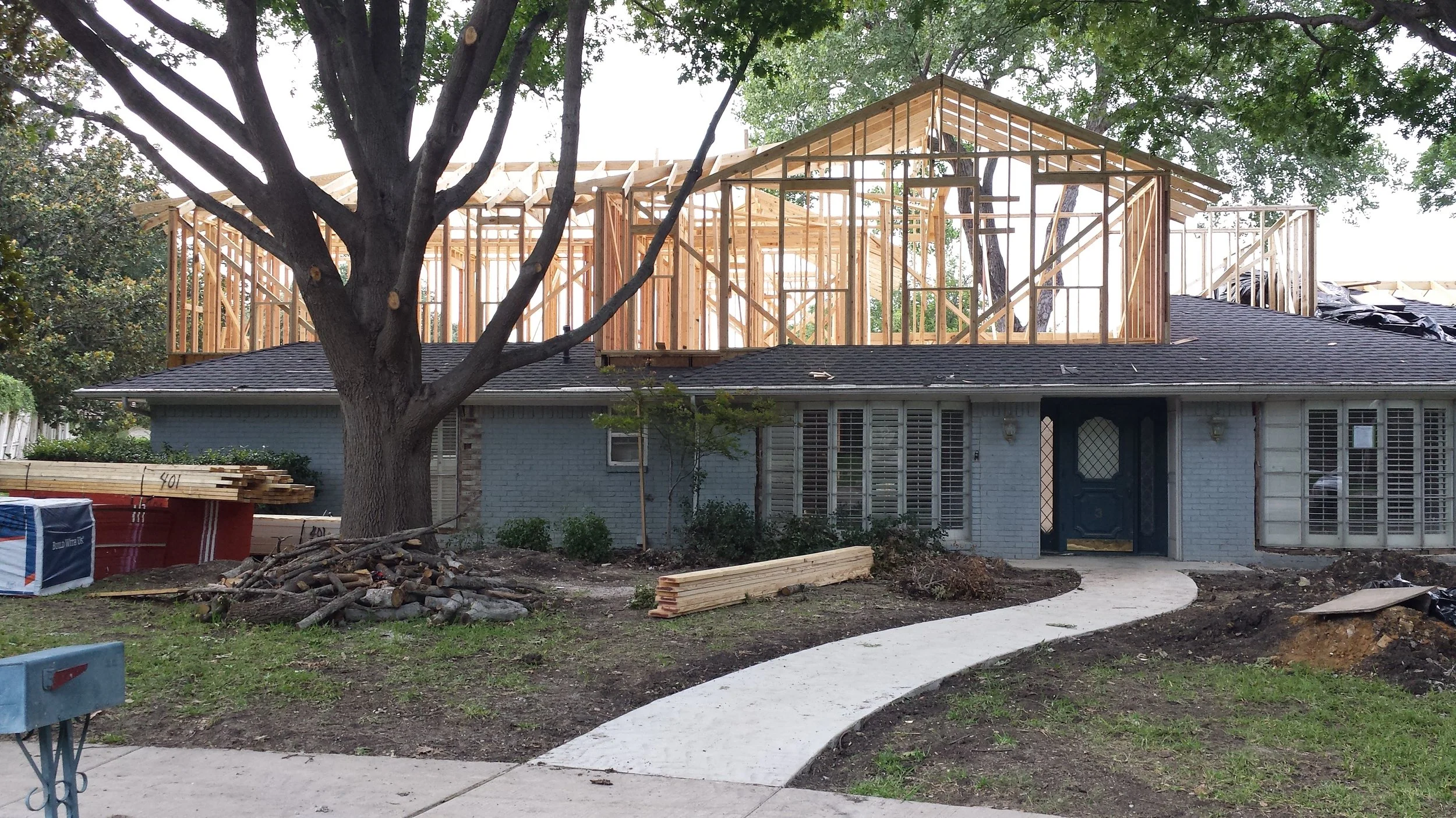 A house under construction with the roof framing in progress. The house has a light blue brick exterior and is surrounded by trees and construction materials.