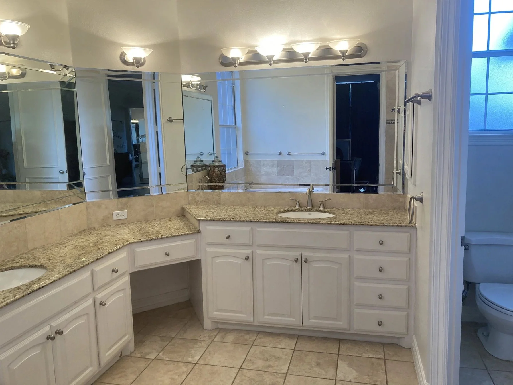 Bathroom with white cabinets, granite countertops, large mirror, and beige tiled floor and walls. There are multiple light fixtures above the mirror and a window on the right side. A toilet is partially visible in a separate room on the right.