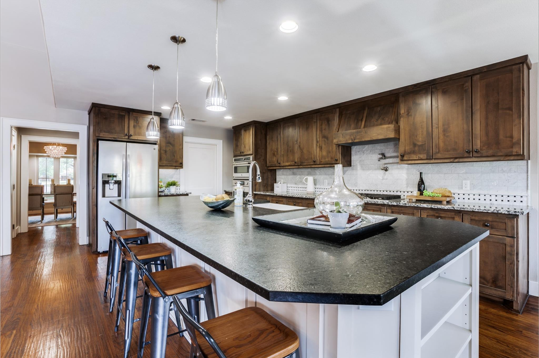 Modern kitchen with large island, dark granite countertops, and wooden cabinets. Stainless steel appliances, pendant lighting, and hardwood floors. Decor includes a glass vase, small potted plant, and bowls.