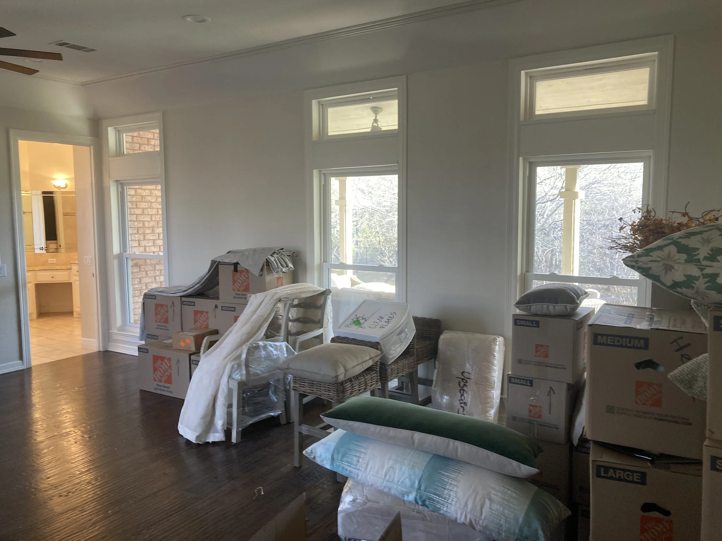 A cluttered living room with moving boxes, pillows, and chairs, near large windows with sunlight streaming in, suggesting a move or renovation.
