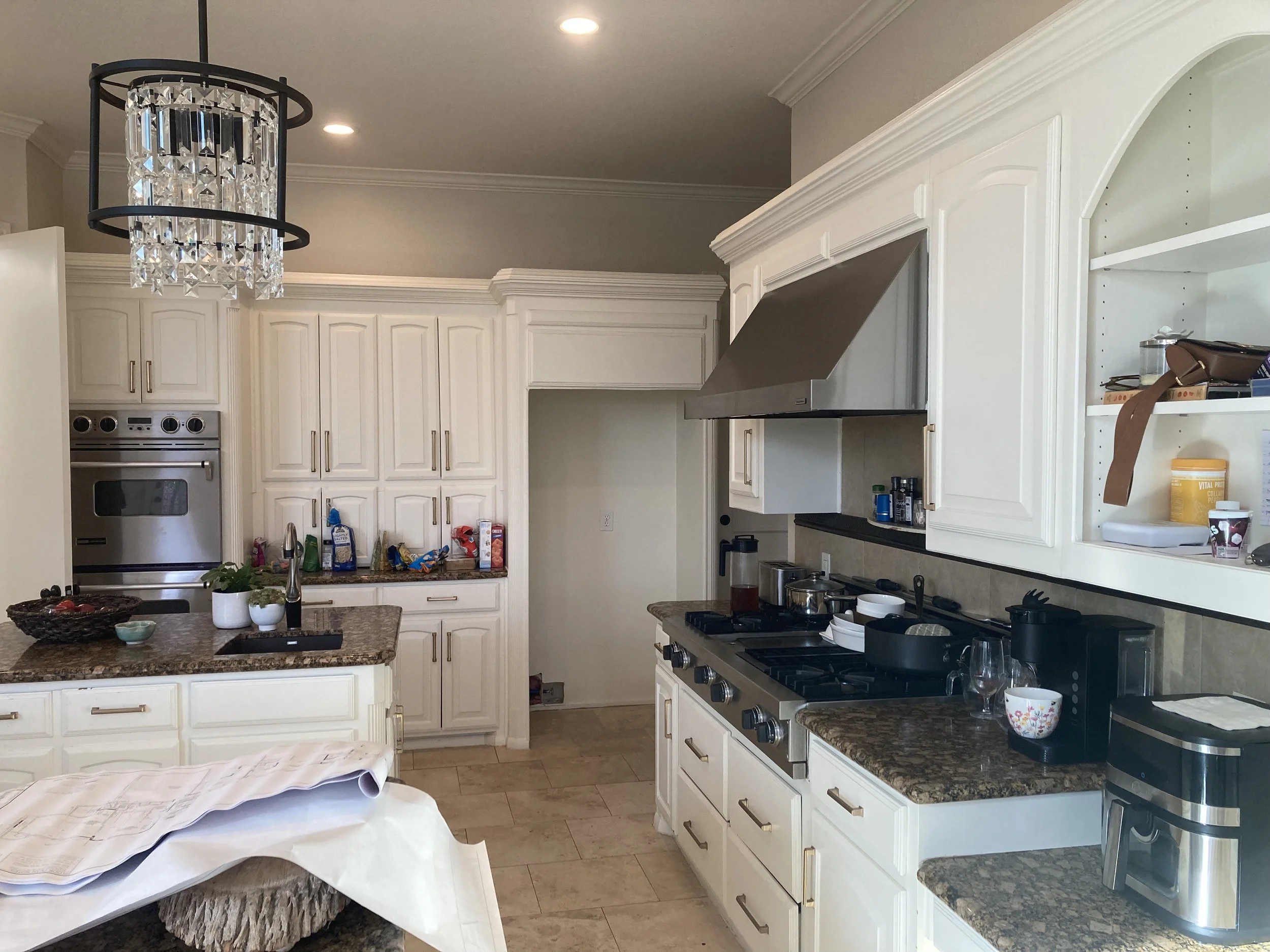 A kitchen with white cabinets, a granite countertop, and stainless steel appliances, including an oven and a range hood. The kitchen is cluttered with various items on the counters and shelves, and a decorative crystal chandelier hangs from the ceiling.