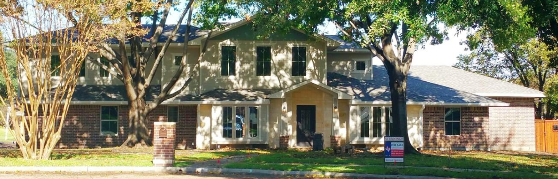 A two-story house with a brick and light-colored exterior, large windows, and a front porch, surrounded by trees with shadows on the house. A 'For Sale' sign is in the yard.