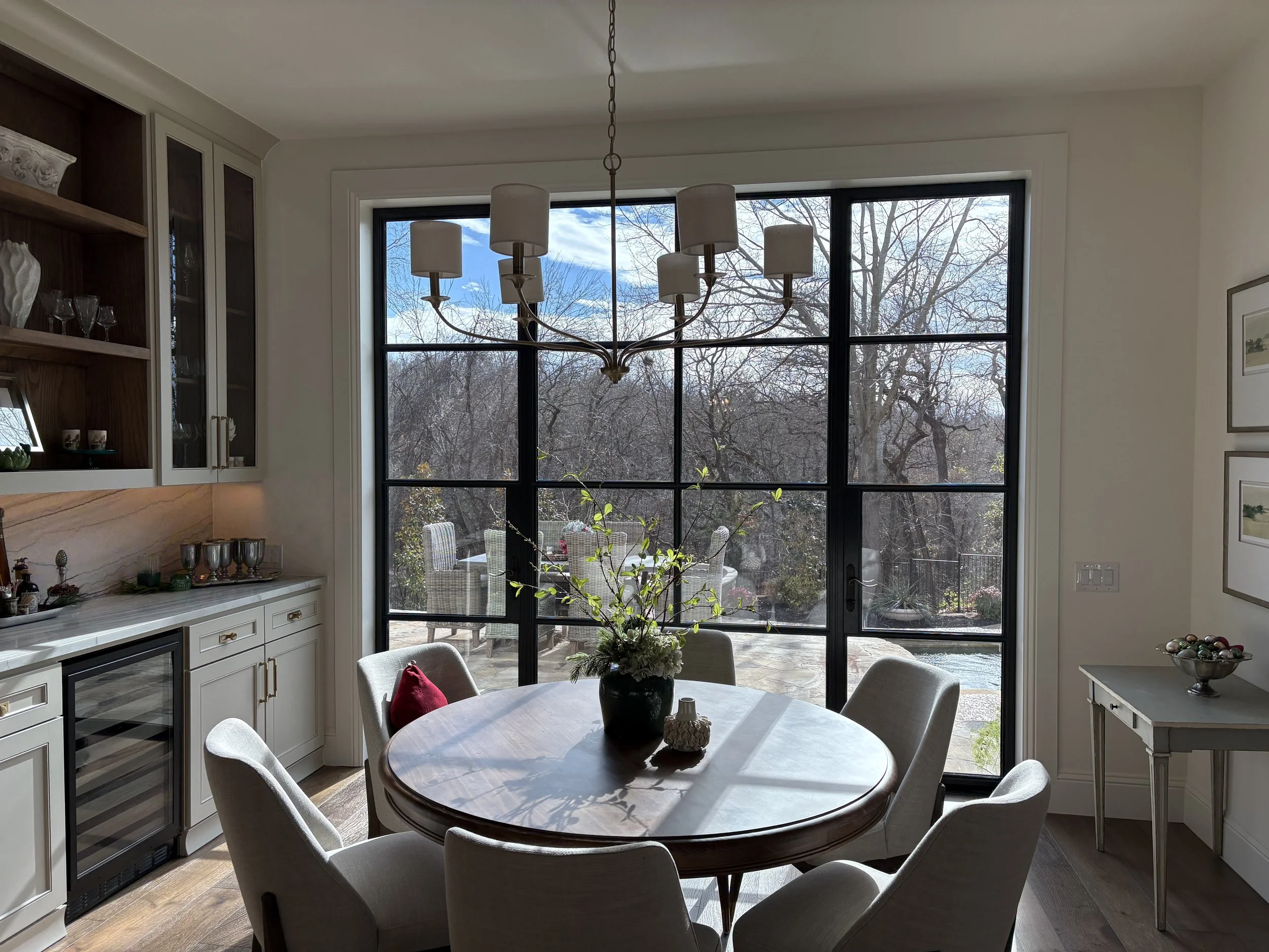 Dining room with large glass window showing trees and sky outside. Round table with a dark vase and plants, surrounded by six gray upholstered chairs. Bright, neutral decor with built-in cabinets and a chandelier.