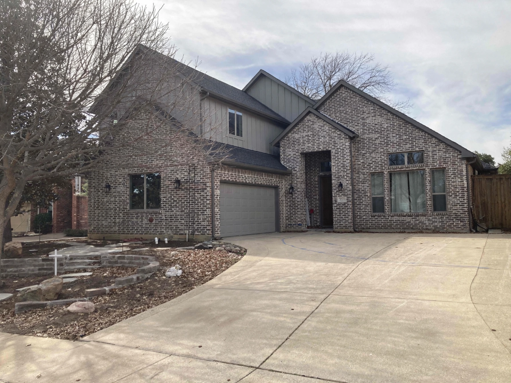 A two-story house with brick and siding exterior, a grey garage door, tree without leaves in the front yard, and a wide driveway.