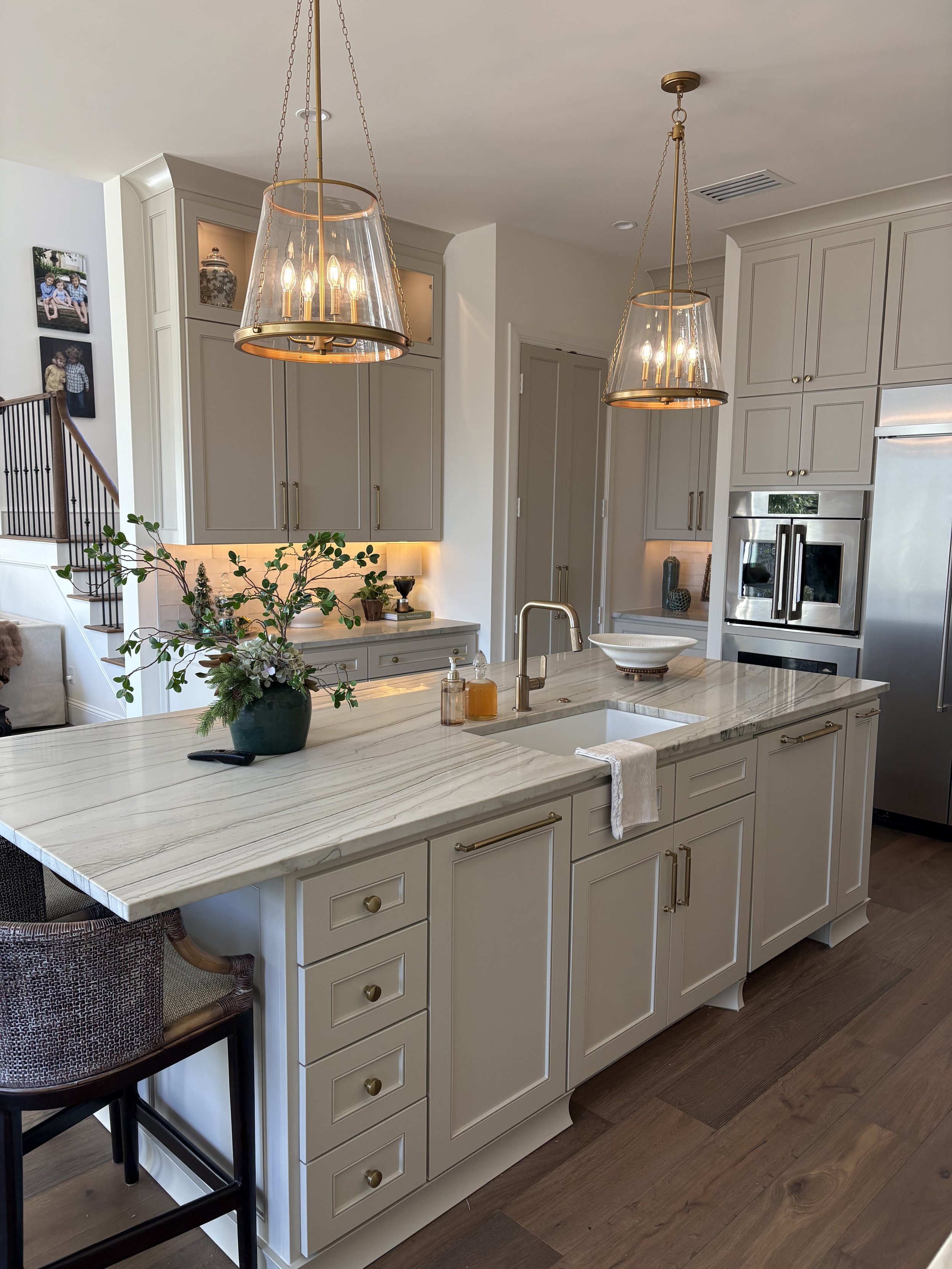 Modern kitchen with white cabinetry, marble island, gold hardware, hanging pendant lights, stainless steel appliances, and decorative plants and photos.