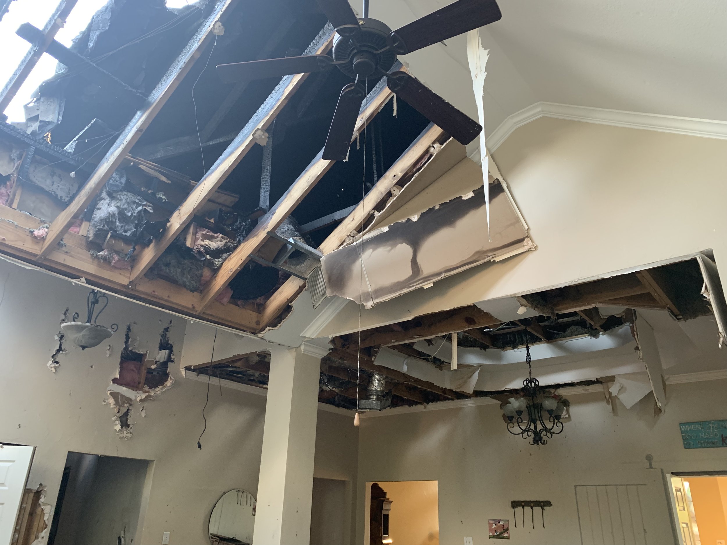 Living room ceiling under renovation with drywall removed, exposing wooden beams, insulation, ductwork, and wires, with a ceiling fan and chandeliers.