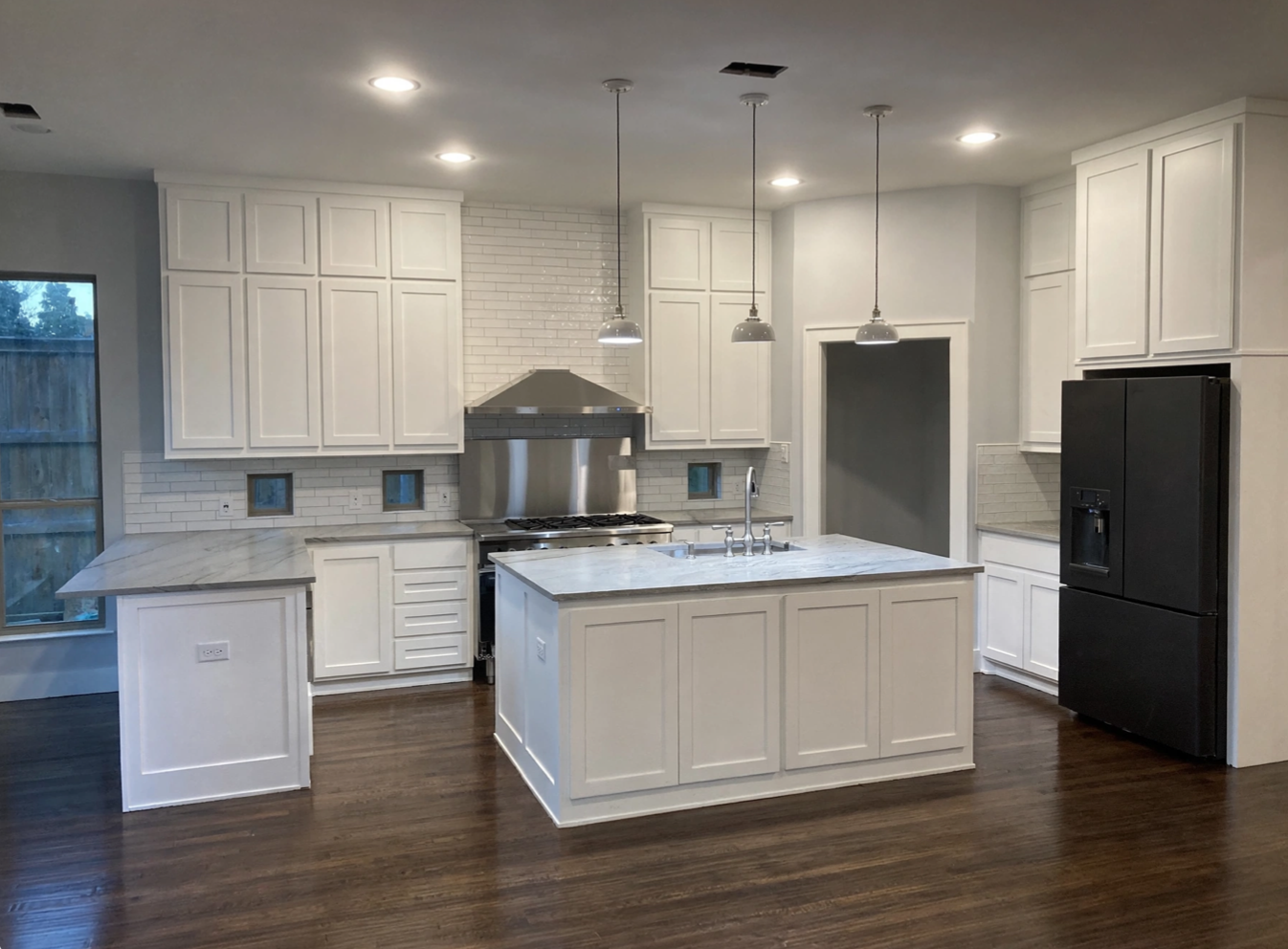 Modern kitchen with white cabinets, marble countertop island, stainless steel stove and range hood, black refrigerator, wood flooring, pendant lights, and a window with a wooden fence outside.