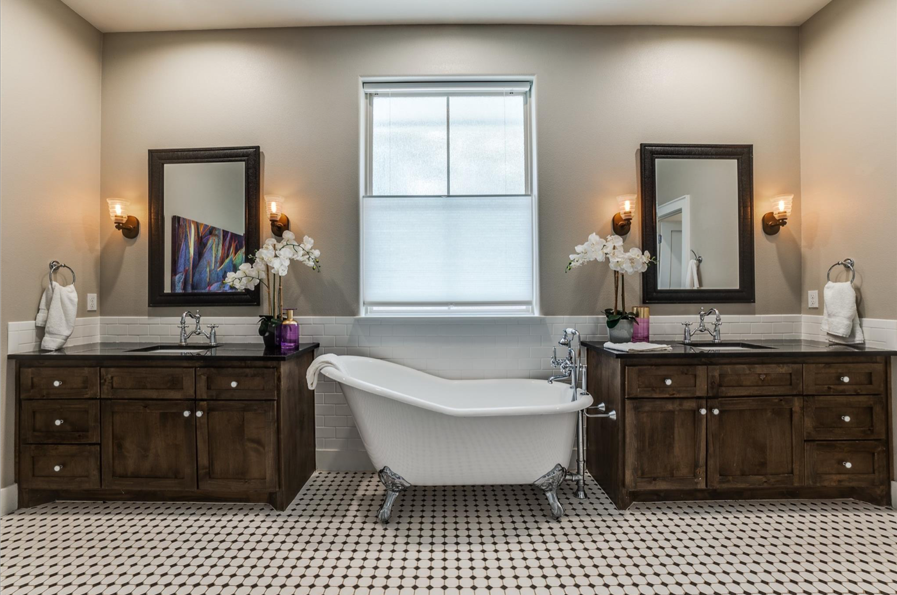 Bathroom with a vintage clawfoot bathtub between two wooden vanities with dark countertops, each with a mirror and light fixture. White subway tile surrounds the bathtub with two orchids on the counters, and a window above the tub. The floor has black and white hexagon tiles.