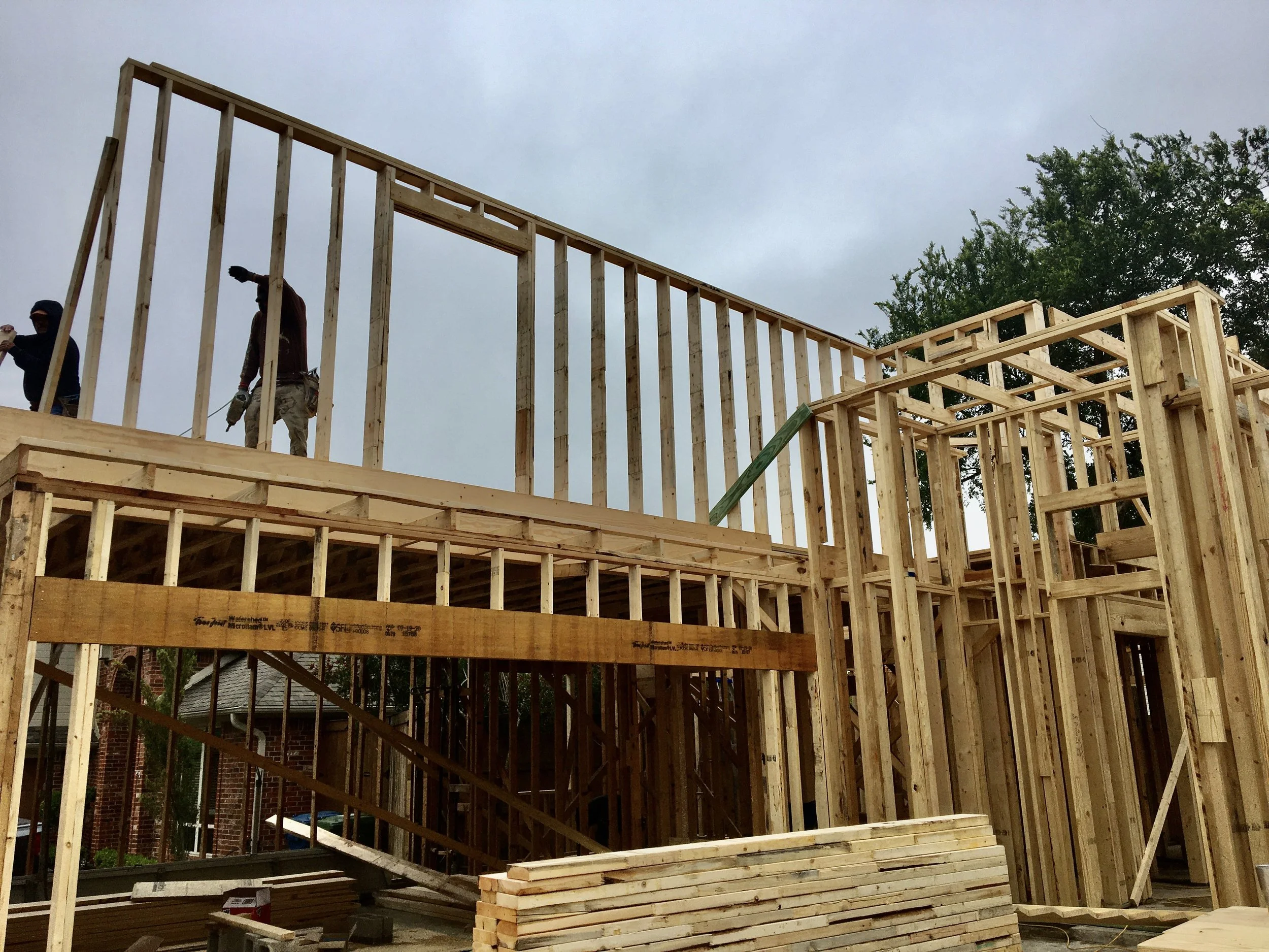 Construction site showing wooden framing of a building under construction with two workers on site, some construction materials on the ground, and a tree in the background.