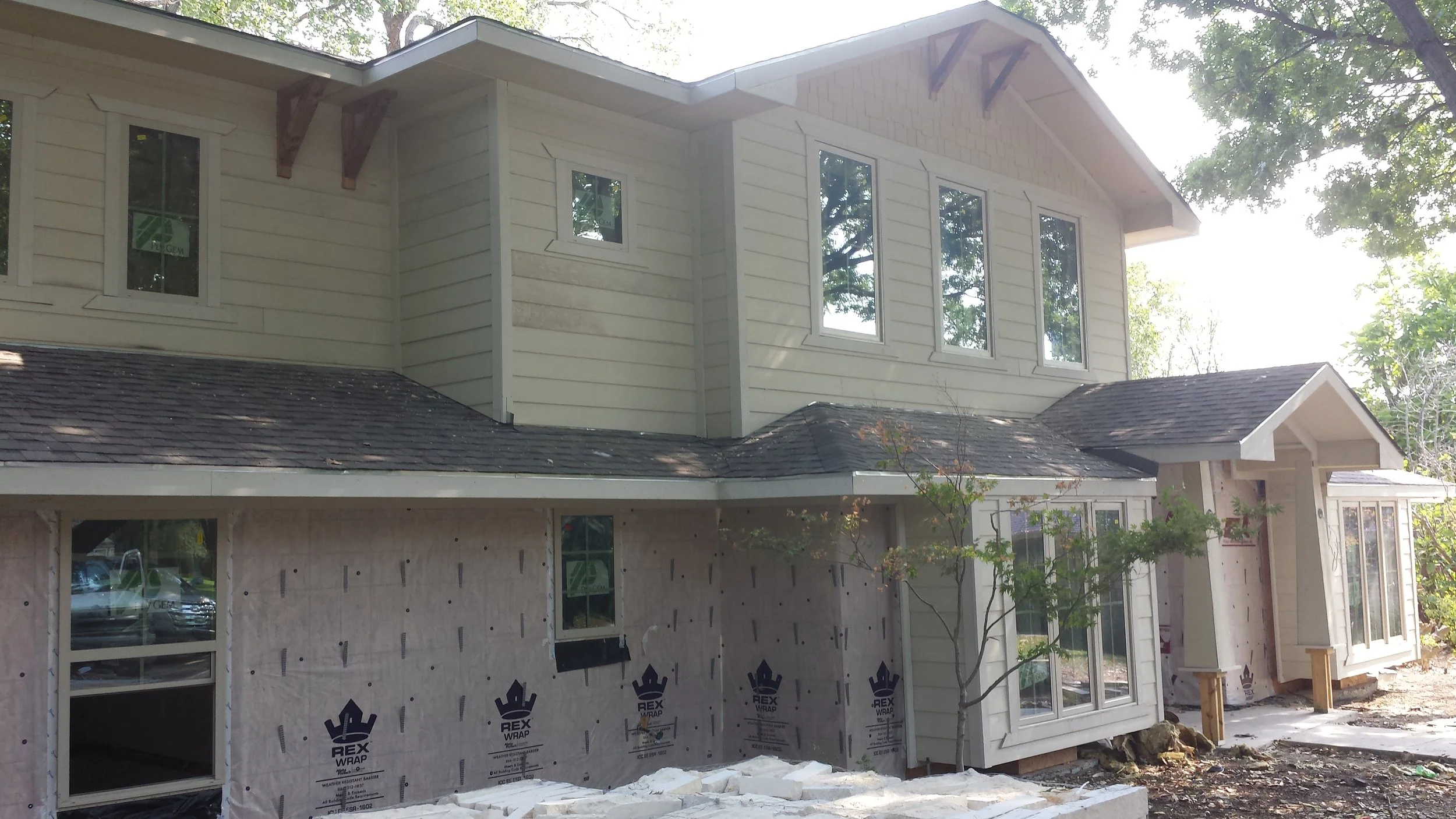 A two-story house under construction with siding partially installed, showing windows and a roof, surrounded by trees.