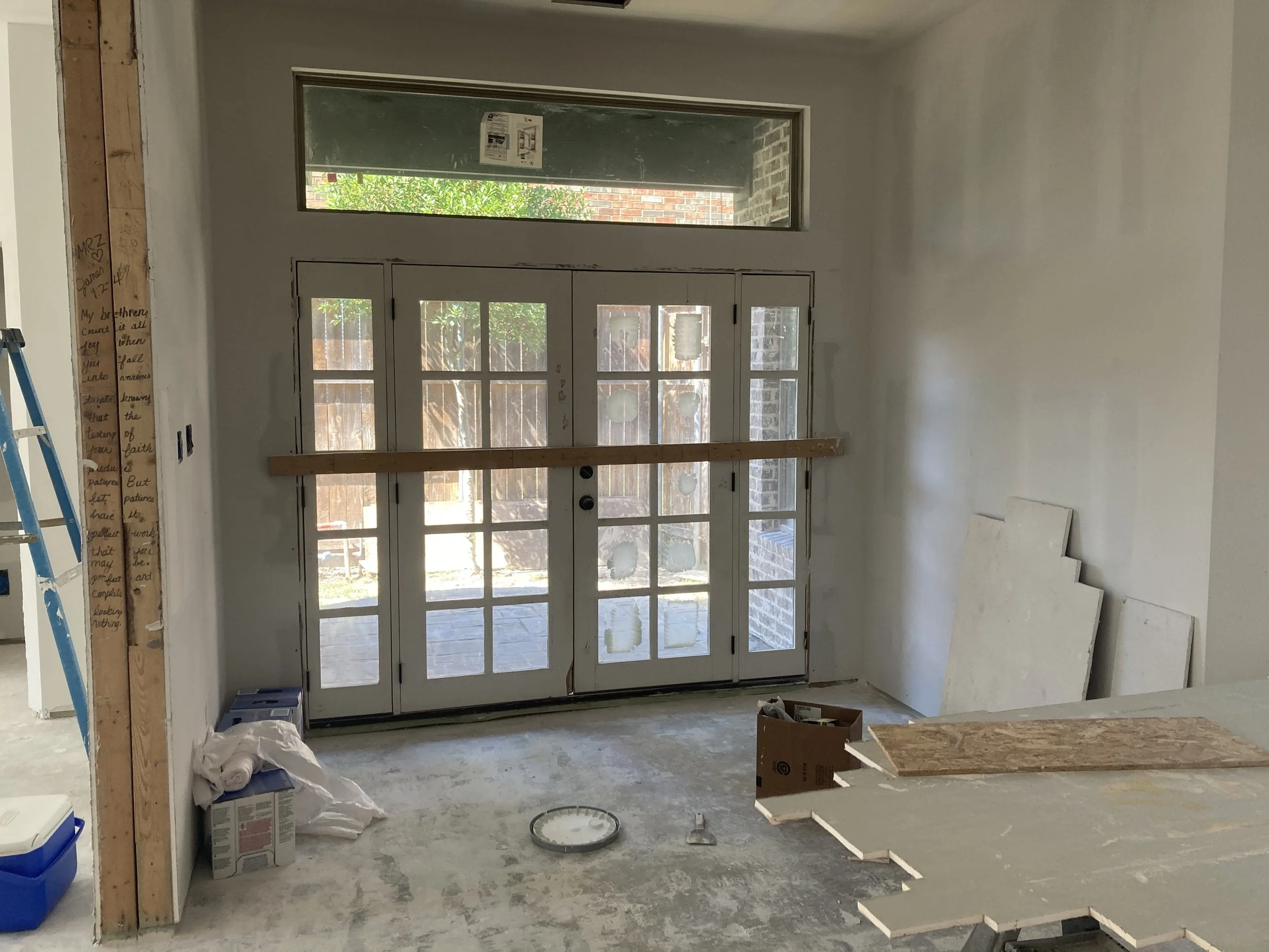 Interior of a house under construction with French doors leading to a backyard, unfinished walls and construction materials on the floor, and a blue ladder on the left side.