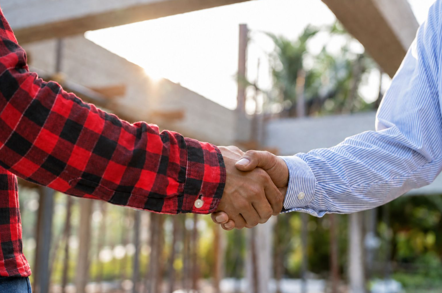 Two people shaking hands outdoors, one wearing a red and black checkered shirt and the other wearing a light blue striped shirt, at a construction site with wooden beams and greenery in the background.