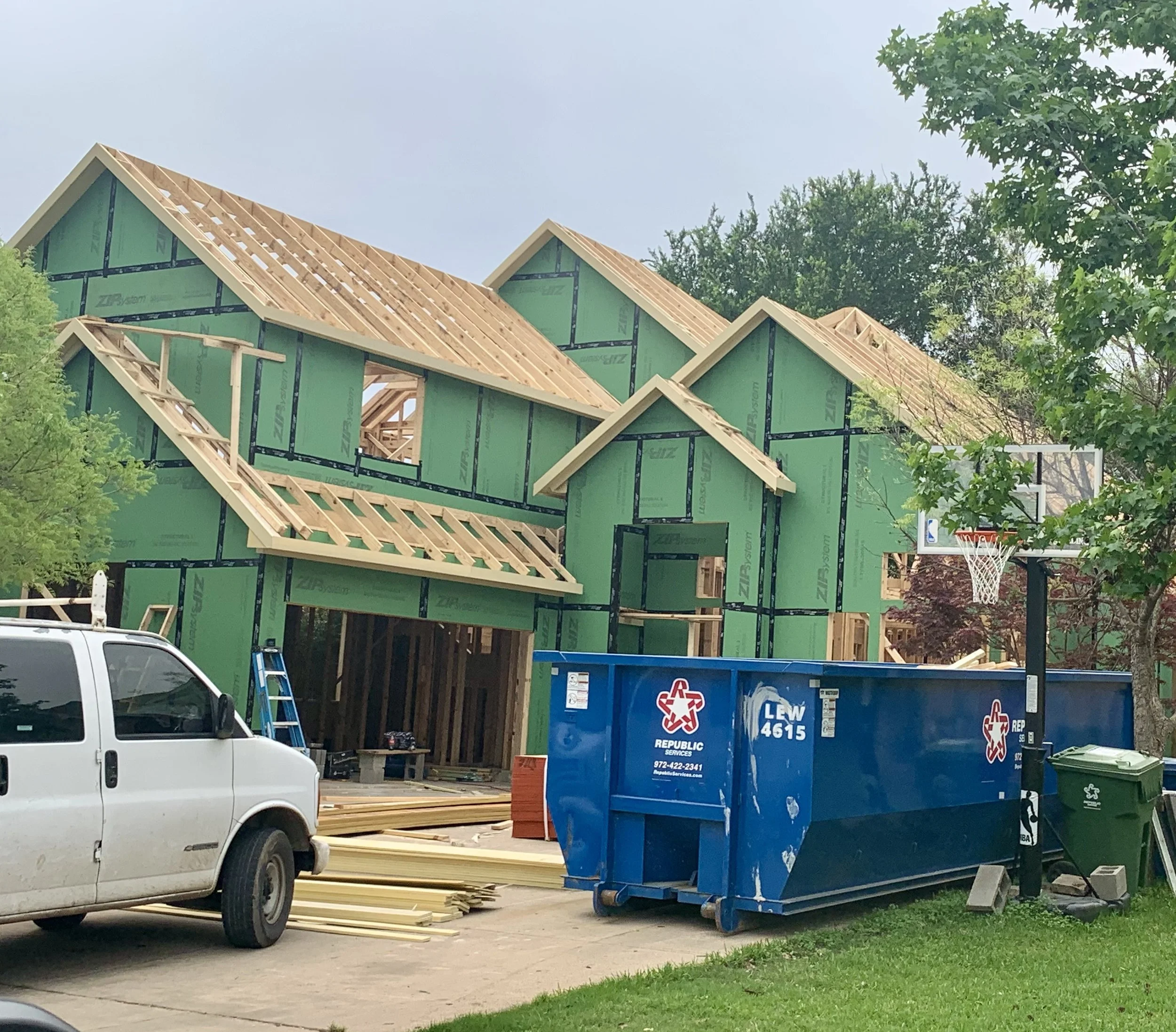 House under construction with wooden roof framing, surrounded by trees, parked vehicle, and a blue trash container.
