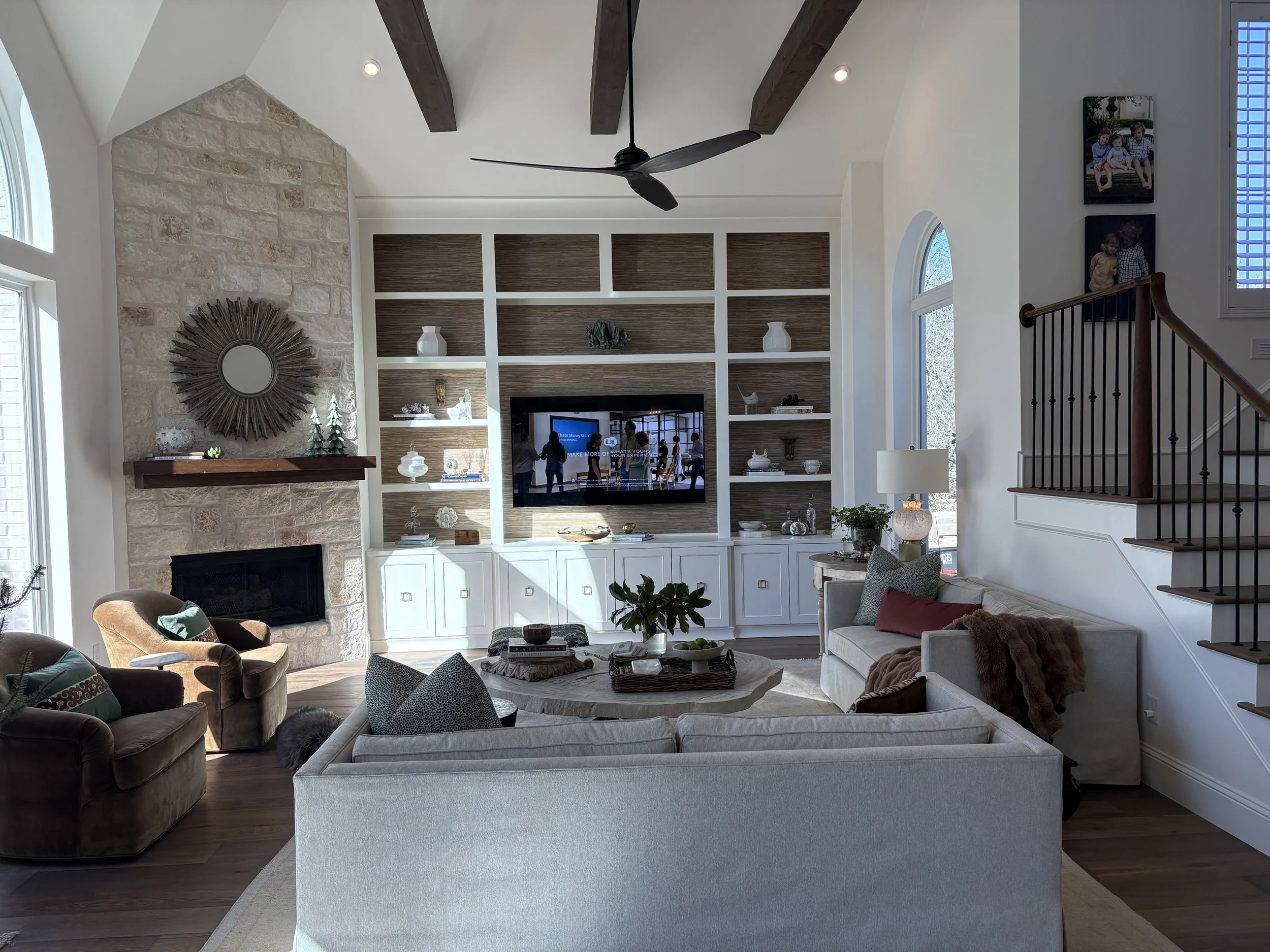 Living room with beige and white sofas, a stone fireplace with a round sunburst mirror, a built-in white bookshelf with decorative items, and a large window with blue shutters. There are wooden beams on the ceiling and a ceiling fan. Family photos hang on the wall near the staircase.