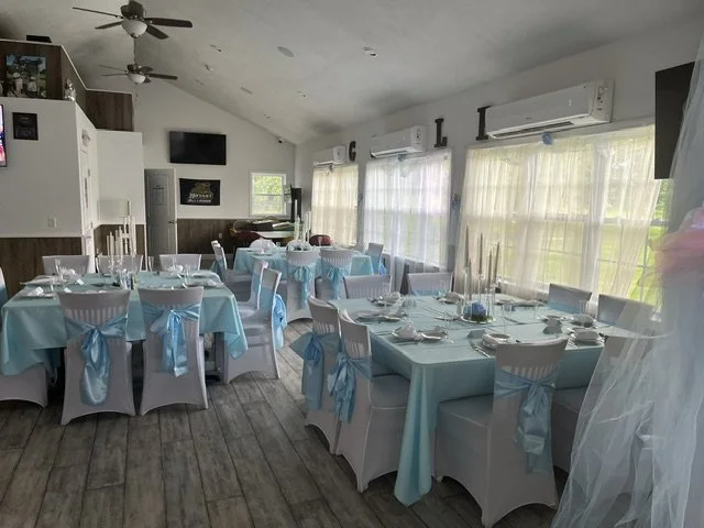 A room decorated for a celebration with round tables covered in light blue tablecloths and chairs with white covers and light blue sashes. The tables are set with plates, silverware, glasses, and candles. The room has large windows with sheer curtain