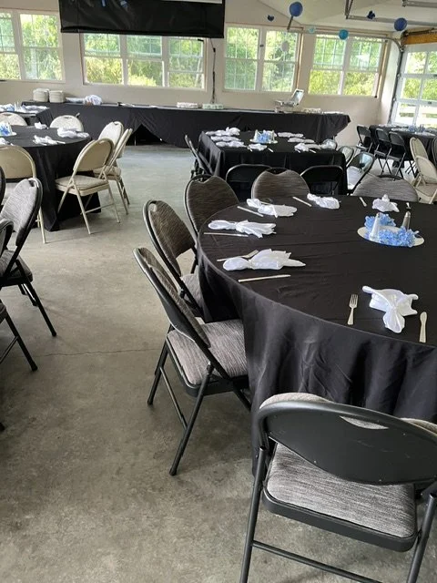 Event venue with round tables covered in black tablecloths, set with white napkins, silverware, and blue centerpieces, with chairs arranged around the tables and windows in the background.