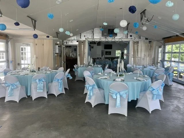 Event space decorated for a celebration with light blue and white theme, round tables with white chairs that have light blue sashes, hanging paper lanterns in blue and white, and large windows letting in natural light.
