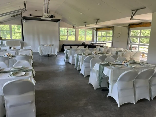A decorated banquet hall with round tables covered in white tablecloths, set with plates, napkins, and centerpieces, ready for a event, with large windows letting in natural light.