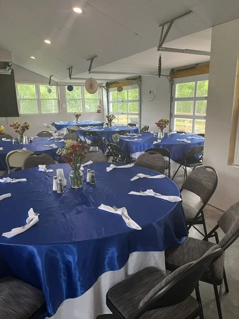 Event room decorated for a gathering with round tables covered in blue tablecloths, each with floral centerpieces, napkins, and salt and pepper shakers, set in a bright room with large windows and hanging paper decorations.