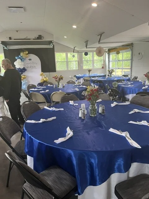 A banquet hall decorated for a celebration with round tables covered in blue tablecloths, set with napkins and silverware, and adorned with floral centerpieces. There are gold and white balloons in the background and a stage with a sign that reads 'H