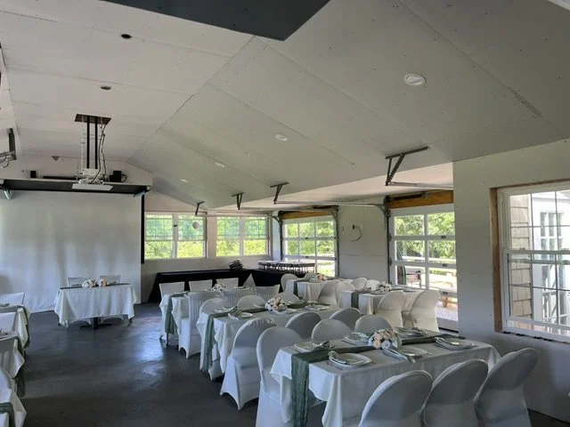 Empty banquet hall with white tablecloths, chairs, and place settings, overlooking a green outdoor area through large windows.