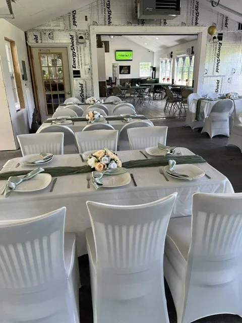 A dining area set up for an event with a long table covered in a white tablecloth, decorated with a green table runner and floral centerpieces. There are white chairs around the table and additional seating in the background, with a television on the