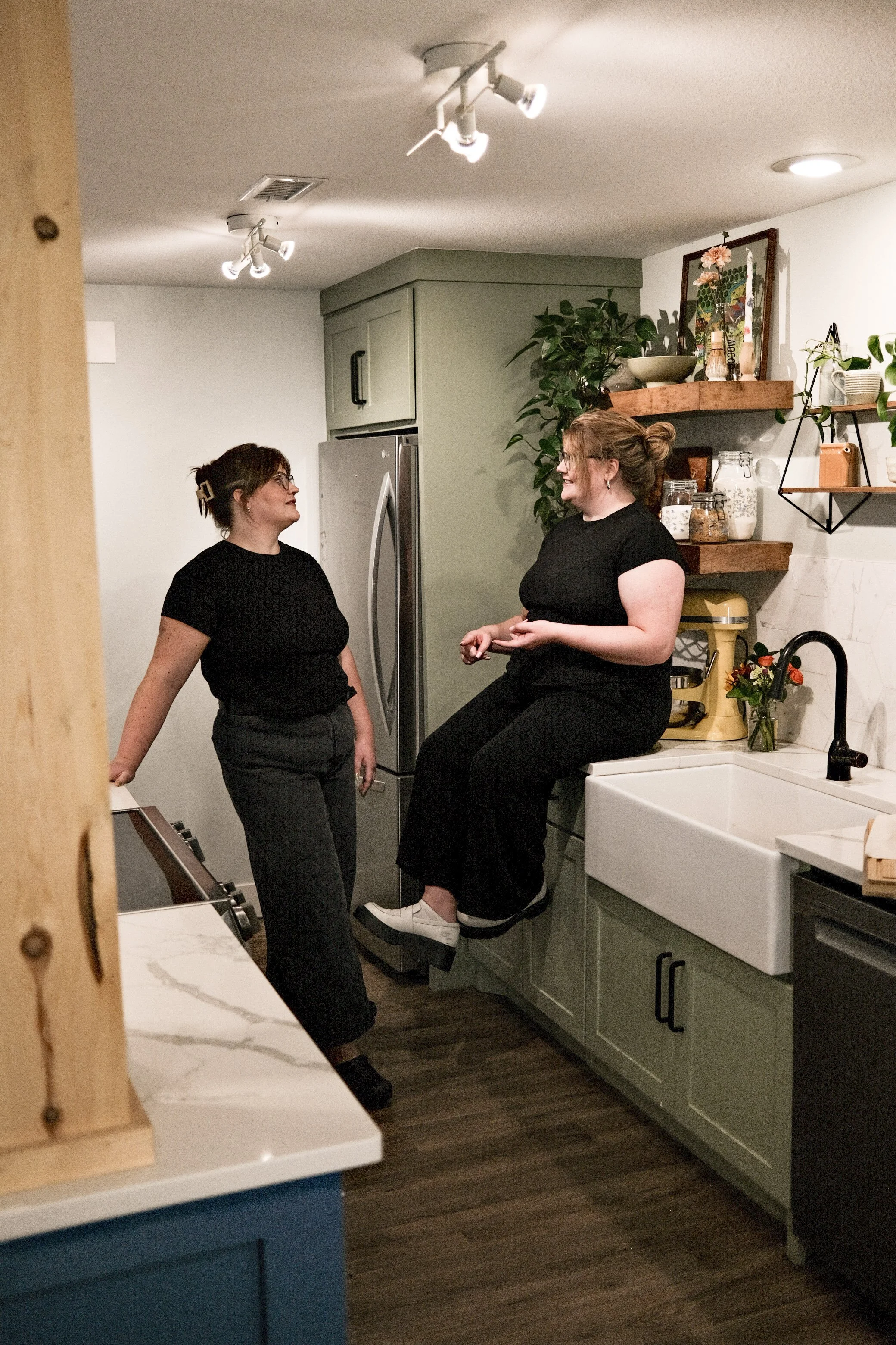 Two women with glasses and black shirts engage in conversation in a kitchen, one sitting on the counter and the other standing beside her.